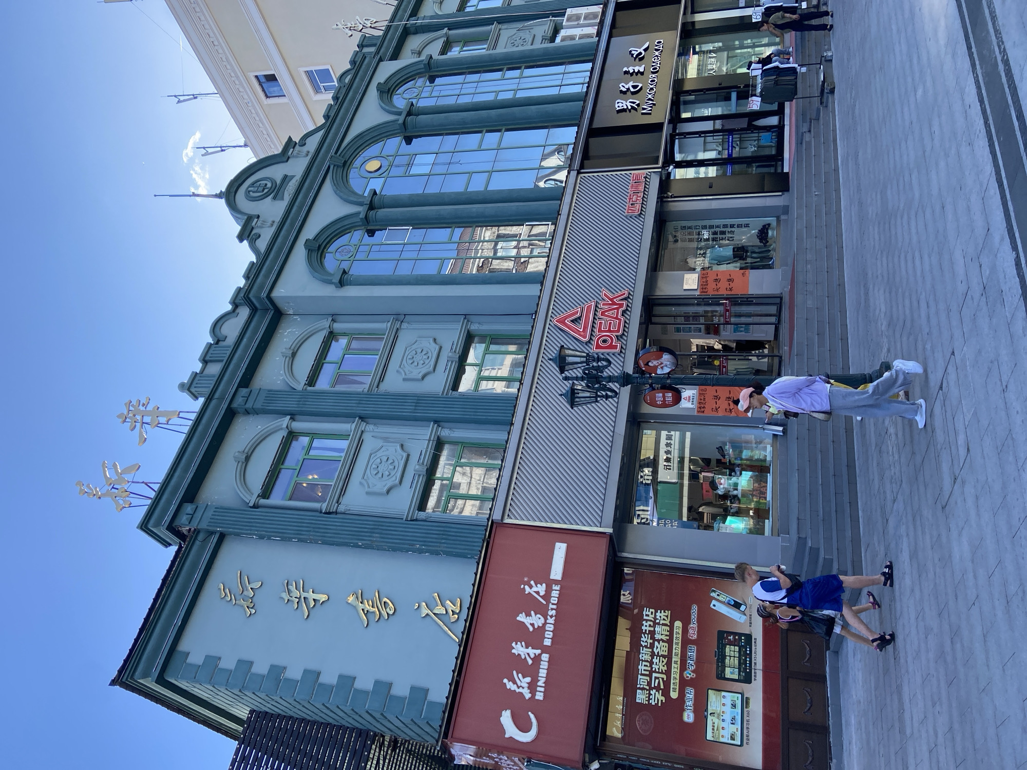 The image depicts a street scene in Xing'an Subdistrict, China. A light blue building with ornate green trim and gold Chinese characters dominates the frame. The building features large arched windows with reflections of other buildings and the sky. Gold lettering is visible on the upper facade. Below the main building, several storefronts are visible. A red sign with white lettering reads "BINHUA BOOKSTORE". To its right, a store with a red triangular logo and "PEAK" in white text is apparent. Further to the right, a store with black signage displays gold Chinese characters and the text "Мужская одежда" (Men's Clothing) in Russian. A lamppost stands in front of the Peak store. People are walking on the paved sidewalk in front of the businesses. Two young people, a male and a female, are walking together towards the right. The male is wearing a blue t-shirt and shorts, and the female is wearing a white top and skirt. Further to the right, a woman in a light purple jacket and grey pants walks towards the left. Another woman in a green jacket and dark pants stands near a rack of clothing outside a store. The sky is clear and blue, with some wispy clouds. The overall scene suggests a commercial district on a clear day.