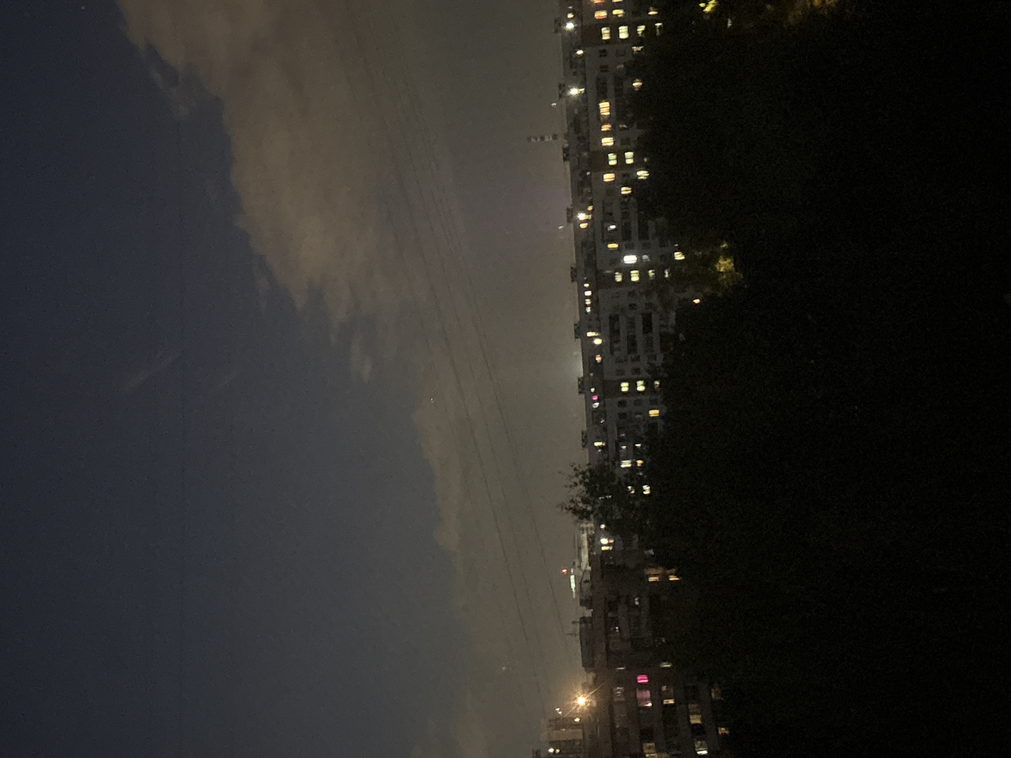 The image, when viewed rotated 90 degrees counter-clockwise, presents a nighttime urban or suburban scene in Moscow, Russia. The sky, occupying the upper portion, transitions from a dark blue-grey on the left to a lighter, hazy grey-brown on the right, suggesting light pollution and low clouds or fog. Several thin power lines traverse the sky horizontally.
Below the sky, a continuous line of multi-story buildings stretches across the middle ground. These structures, likely residential or commercial, are dotted with numerous illuminated windows, displaying a mix of warm yellow, orange, and some cooler white, pink, or red lights. The buildings appear somewhat distant and partly obscured by the atmospheric haze, particularly towards the right side of the frame. A diffuse, bright light source, possibly the moon or a significant artificial light, is faintly visible above the buildings in the hazy section. Additional street-level lights are noticeable at the base of some buildings.
The right side of the foreground is dominated by a dark mass of dense foliage, likely trees, which partially obscures the lower parts of the buildings. The scene conveys the quiet activity of a city at night. No people or specific events are visible, and there is no discernible text. The weather appears cloudy or hazy.