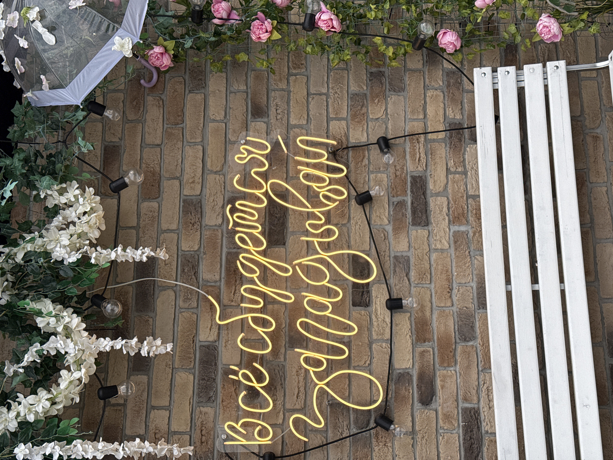 The image shows a close-up of a brick wall with a neon sign that reads "Всё будет хорошо" in yellow neon script. The sign is illuminated and hangs on the wall. To the left of the sign, a string of white artificial flowers and greenery hangs down. To the right of the sign, there is more greenery and pink artificial flowers, along with a clear umbrella decorated with artificial flowers. Several black string lights with exposed bulbs are visible, some hanging above the sign and others to its sides. In the foreground, a white slatted bench is partially visible at the bottom of the frame. The overall setting appears to be outdoors, possibly a cafe or an event space. There are no people in the image.