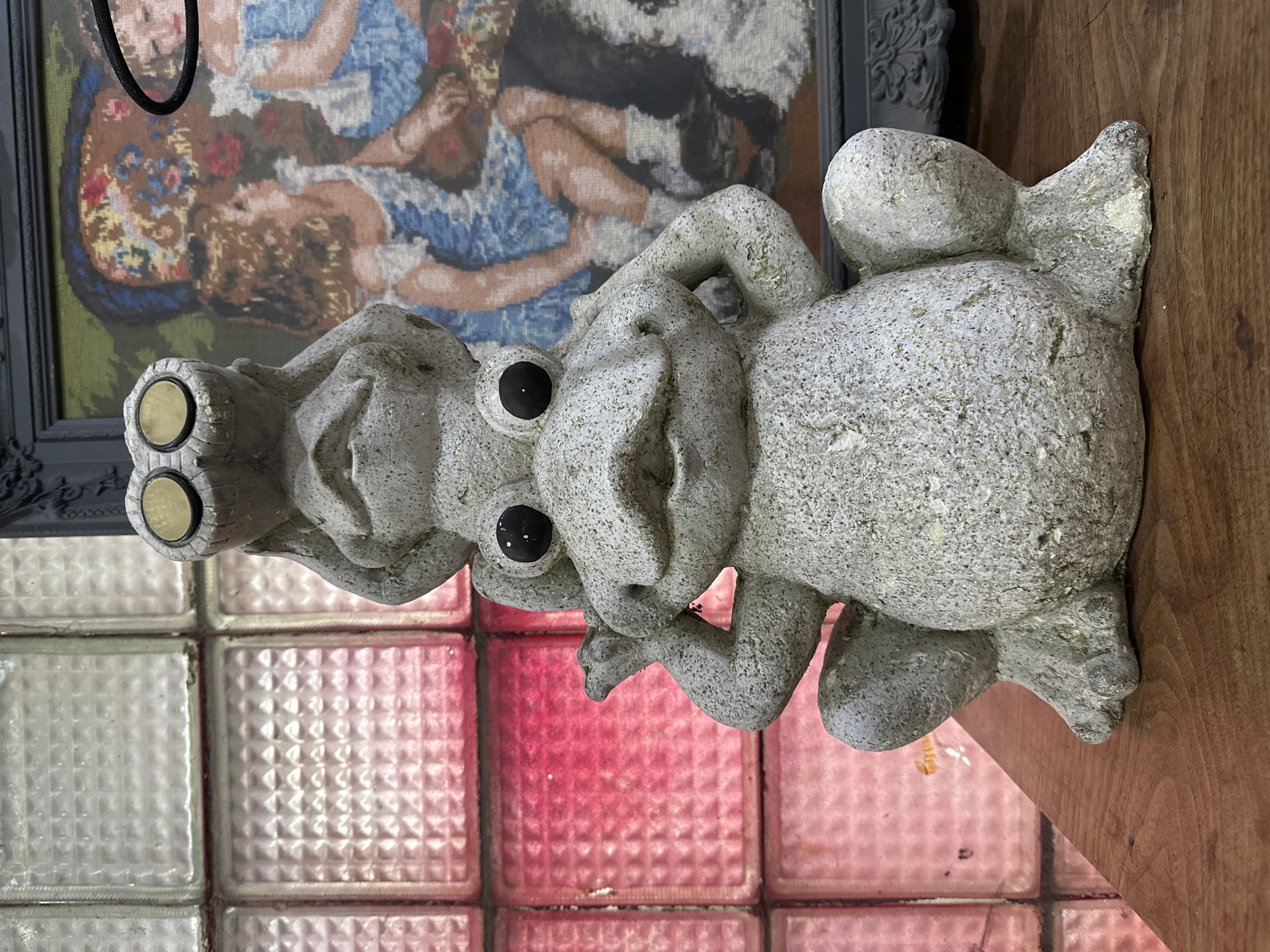 A stone statue of three frogs stacked on top of each other sits on a wooden surface. The frogs are arranged in a "hear no evil, see no evil, speak no evil" motif. Behind the statue is a wall of patterned glass blocks, some tinted pink. A framed tapestry hangs in the background above the statue. The location may be Tbilisi, Georgia, but there are no identifying indicators in the scene.