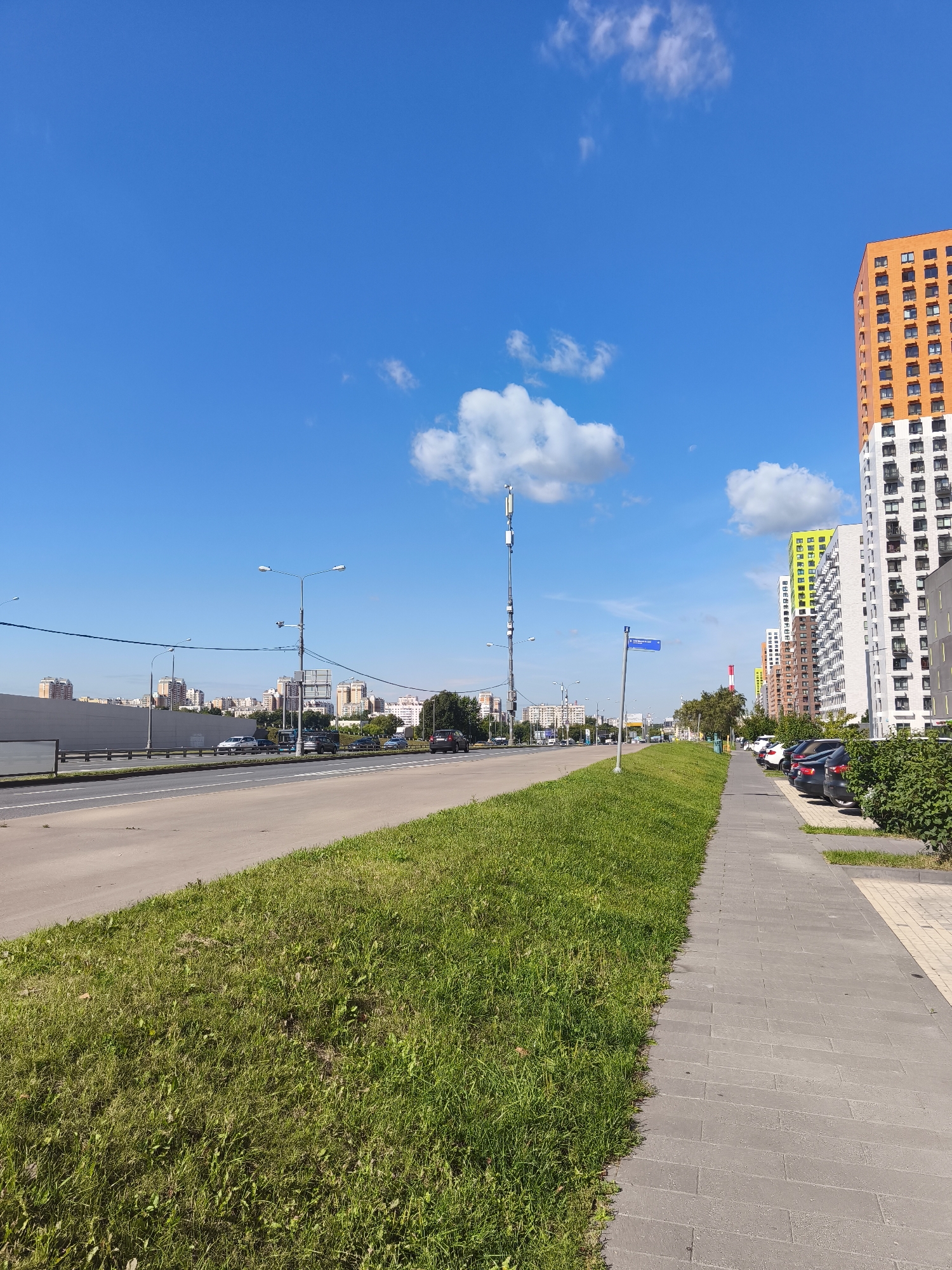 A street scene in a city on a clear day. The foreground features a well-maintained green lawn bordering a paved pedestrian walkway. Beyond the walkway, a multi-lane road carries a light flow of traffic. In the background, modern, multi-story residential buildings with colorful facades (orange, green, white) are visible, set against a bright blue sky with scattered white clouds. A cell tower stands tall amidst the cityscape, and a street sign is visible on a pole. The scene appears to be a residential or mixed-use urban area.