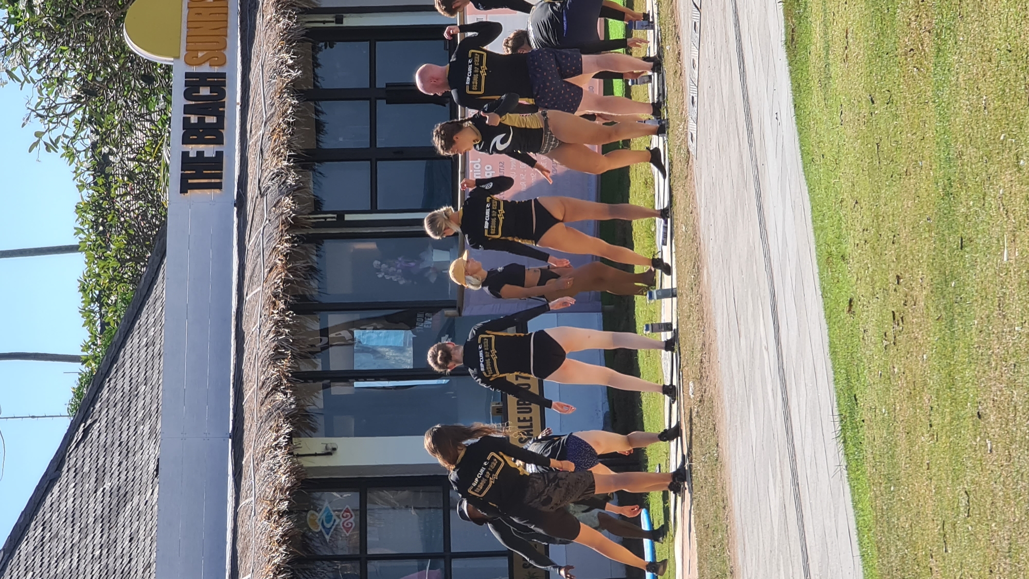 In Denpasar, Indonesia, a group of individuals, predominantly women, are engaged in what appears to be a surfing lesson or a related training activity outside "The Beach Sunrise" establishment. The building features a thatched roof and large windows. The individuals are dressed in swimwear, with many wearing black long-sleeved rash guards featuring "Rip Curl School of Surf" branding. They are positioned on a paved area in front of the building, with some standing and others in various athletic poses, suggesting active participation. Water bottles are visible on the ground near some of the participants. The environment is sunny and outdoors, with grass and trees visible in the background.