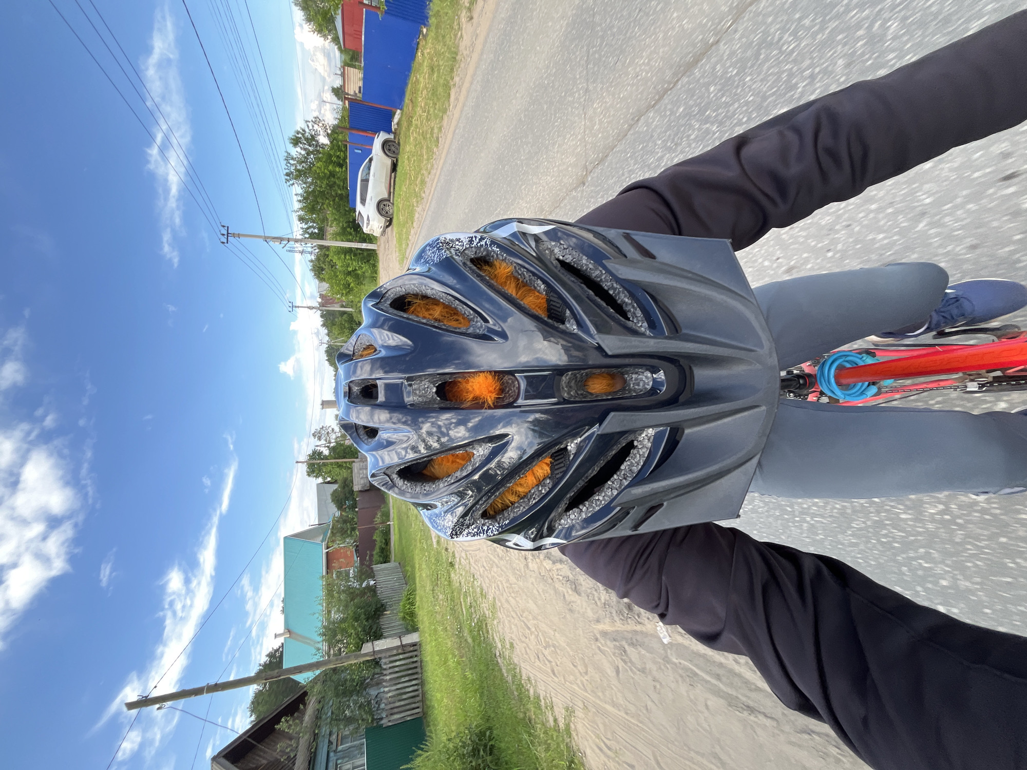 This is a first-person perspective video of someone riding a bicycle in Chaikovsky, Russia. The rider is wearing a black helmet with orange padding visible through the vents, a black long-sleeved shirt, and grey pants. The bicycle's red frame and blue cable lock are visible. The scene is a rural or suburban road with a dirt or gravel shoulder.
On the left side of the road, there are houses with wooden fences and a distinctive teal-colored roof. Further down the road, a white car is parked. The background features a clear blue sky with scattered white clouds. Power lines run parallel to the road, supported by wooden poles, indicating a residential area. The vegetation appears green and lush, suggesting it's a warm day.
The time of day appears to be daytime, likely late morning or afternoon, given the bright sunlight and clear skies. The weather seems pleasant and suitable for cycling. The overall mood is active and perhaps a bit adventurous, as suggested by the point-of-view shot. There is no visible text in the media file.