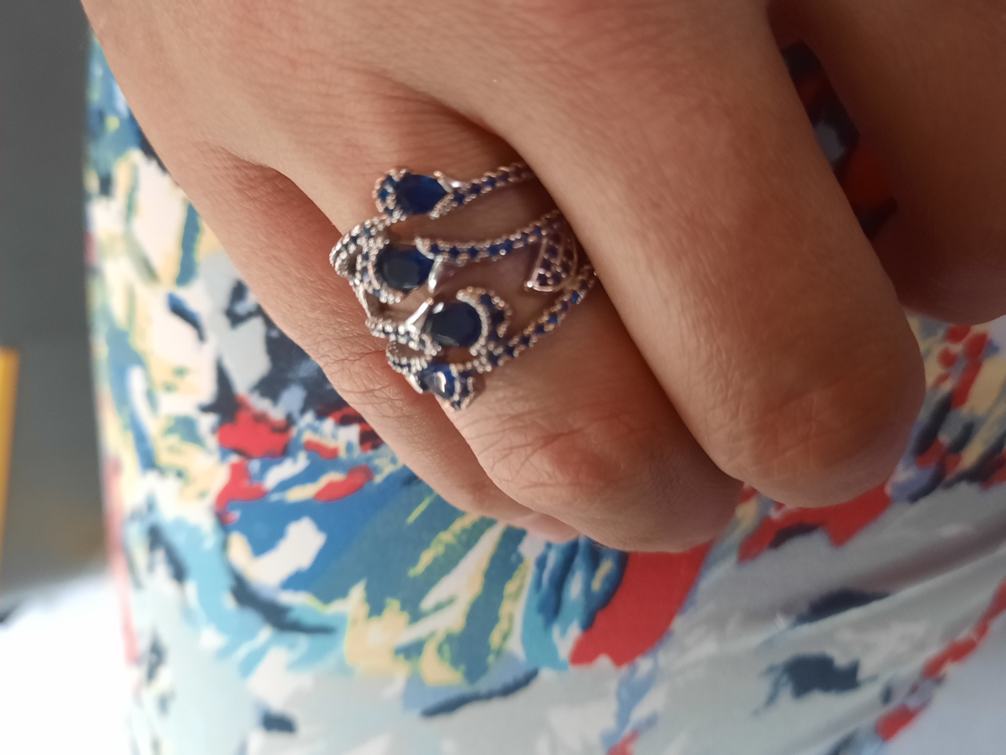 A close-up, high-angle shot captures a hand adorned with an intricate silver ring featuring multiple teardrop-shaped blue gemstones. The ring is positioned on the index finger, with its design extending across the top of the finger. The hand is resting on a surface covered with a colorful fabric that exhibits a vibrant abstract pattern of blues, reds, and yellows. The lighting suggests an indoor environment, possibly a home or studio, with no specific landmarks visible to indicate a location in Rosenheim, Germany.