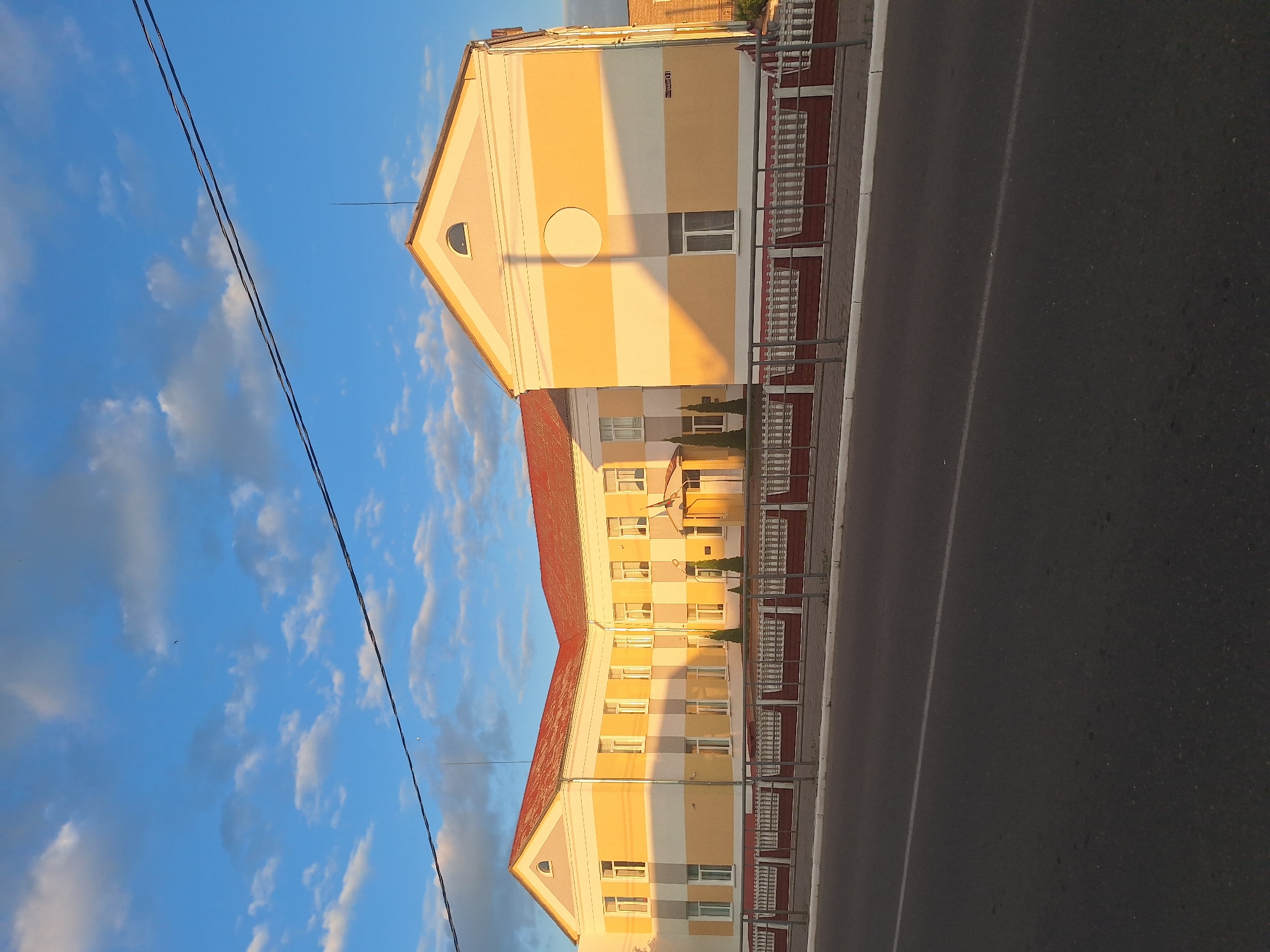 An exterior shot captures a two-story building with a red tile roof and horizontal yellow and white stripes on its facade. The building has numerous windows. A white picket fence with a red trim separates the building from a paved road in the foreground. The sky above is blue with scattered white clouds, and the sun casts a shadow on the building's right side. The overall scene appears to be in a quiet, possibly rural or suburban area. The image is taken at an angle, looking towards the building.