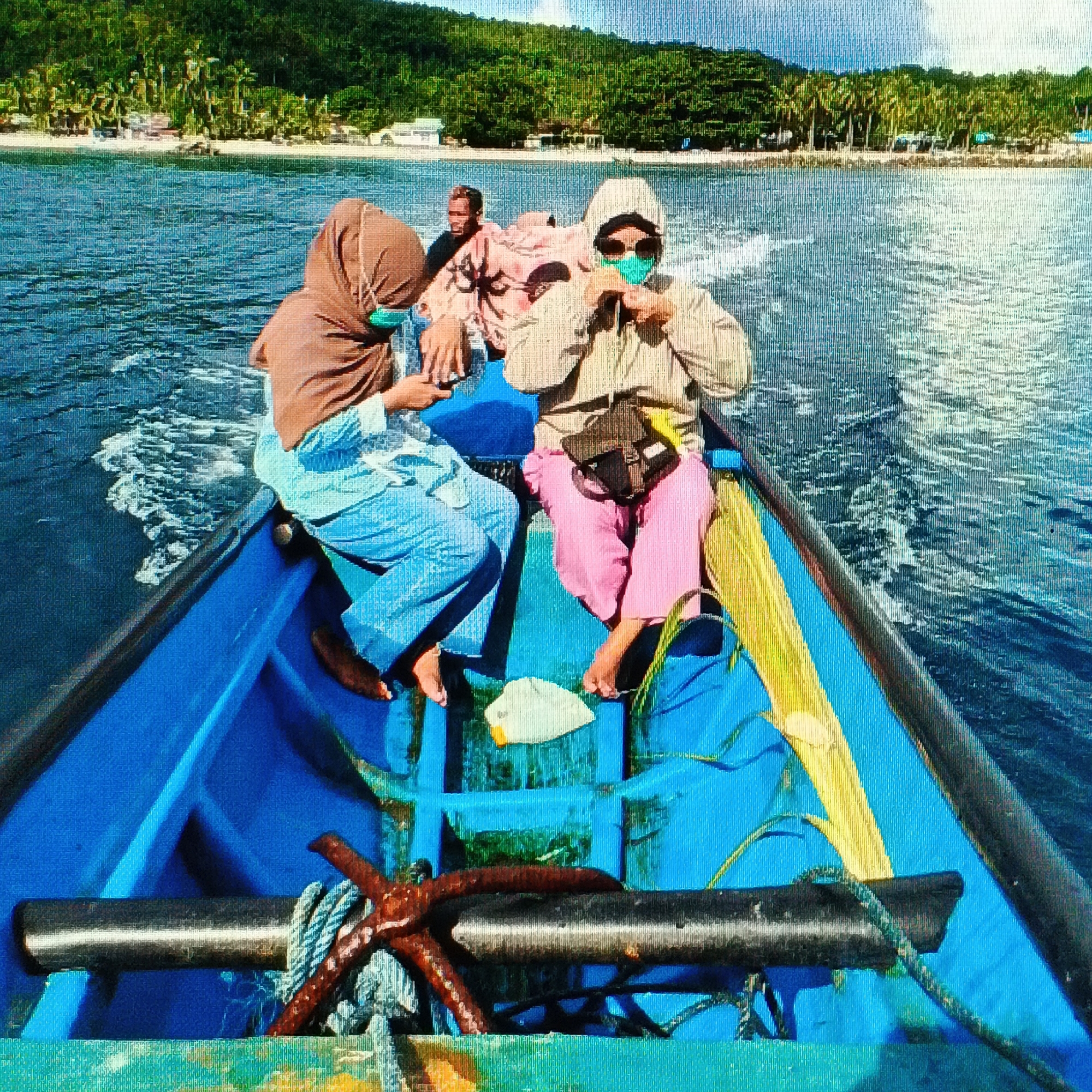 The image depicts three individuals traveling on a blue boat through a body of water, with a lush green, tree-covered island in the background. Two women are seated in the foreground, both wearing face masks. The woman on the left wears a brown hijab and blue pants. The woman on the right wears a light-colored hooded jacket, sunglasses, a face mask, and pink pants. A man is visible in the background, also wearing a mask. The boat appears to be moving, evidenced by the wake in the water. The environment suggests a tropical or coastal setting. The specific location in Ende, Indonesia, cannot be determined from the visual information alone.
