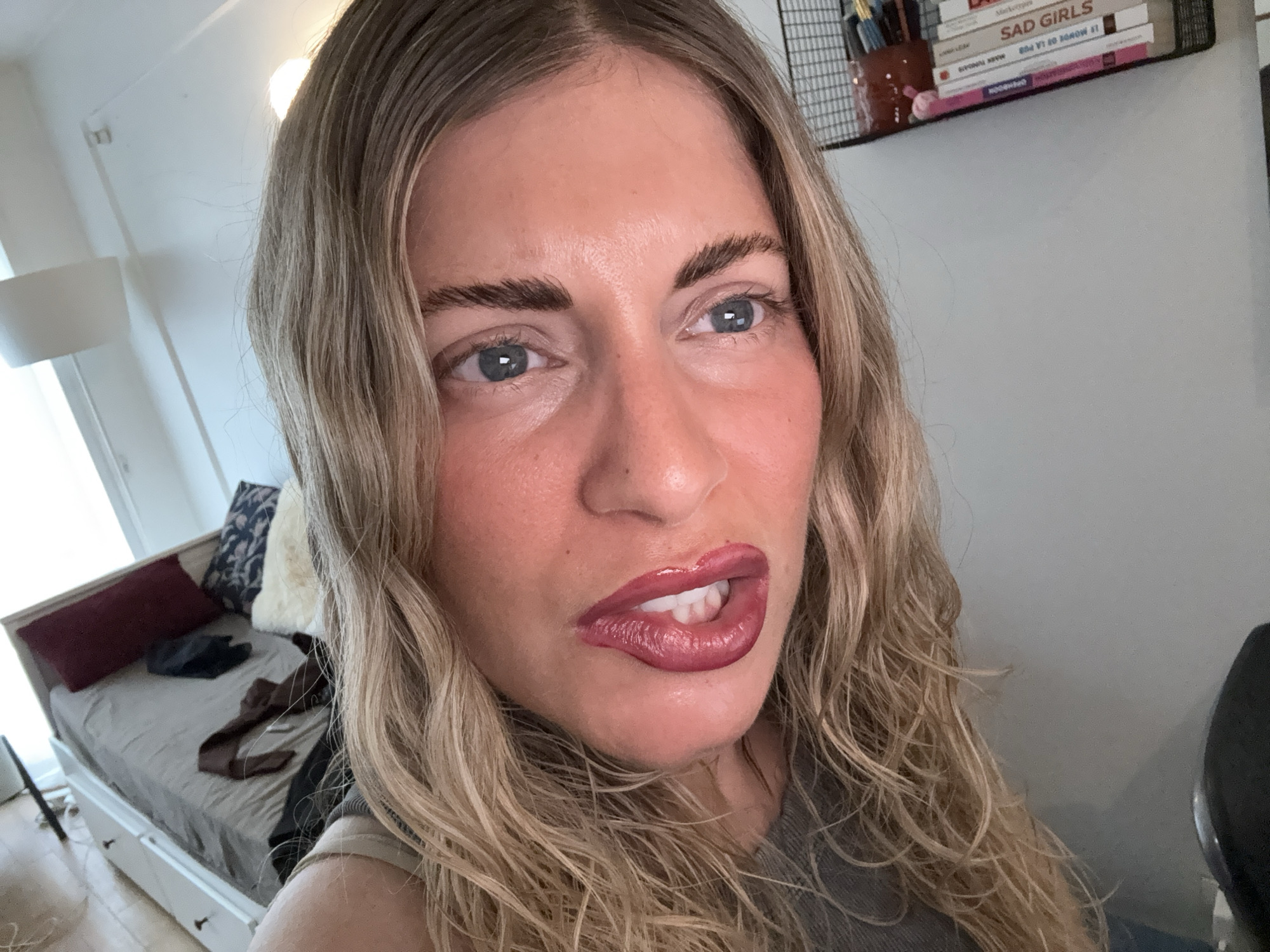 A blonde woman is captured in a close-up, selfie-style shot indoors. Her face is the primary focus, with her blonde hair framing her features. She is wearing dark lipstick and appears to be biting her lower lip. In the background, a bed with pillows and blankets is visible, along with a shelf holding books and other items. The lighting suggests an indoor setting, possibly a bedroom or living space. There is no indication of the location being Paris, France.