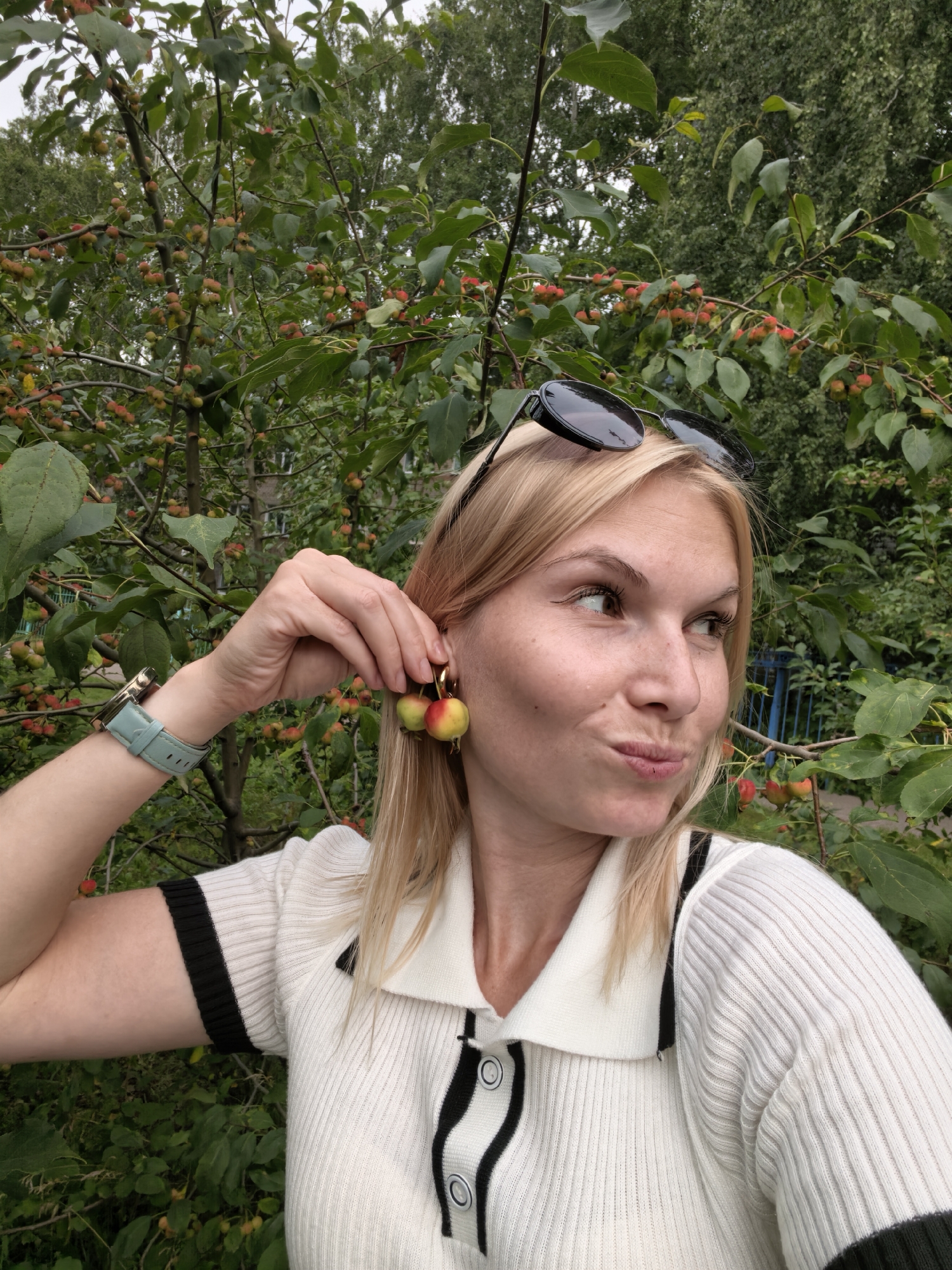 A blonde woman in a white and black collared shirt is holding small, round, red and yellow fruit earrings up to her ear. She is wearing a light blue watch on her left wrist. Sunglasses are perched on top of her head. She is outdoors in front of a tree with small, red and yellow fruits and green leaves. The background is blurry and shows more greenery and a blue fence. The location is the city of Sterlitamak, Russia.