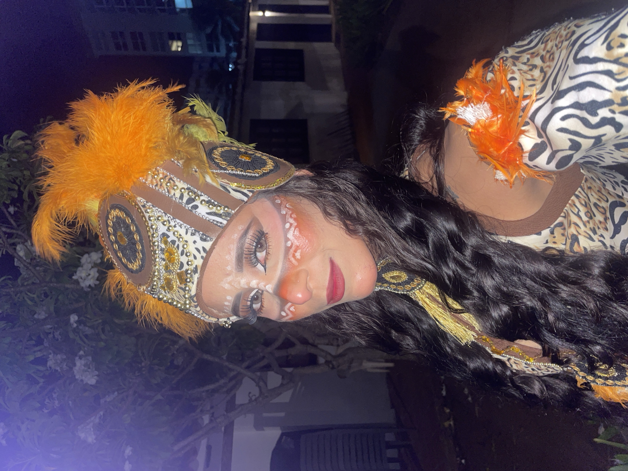 A woman in elaborate tribal-inspired costume and makeup poses for a portrait outdoors at night. She wears a headdress adorned with orange feathers and gold embellishments. Her face is painted with white tribal markings and orange blush. She has long, dark, wavy hair. Her outfit features a leopard print and zebra print pattern, with additional gold and orange feather accents on the shoulder. The background shows a dimly lit outdoor area with a tree to the left and a building with windows in the distance, suggesting a residential or commercial area in Porlamar, Venezuela.