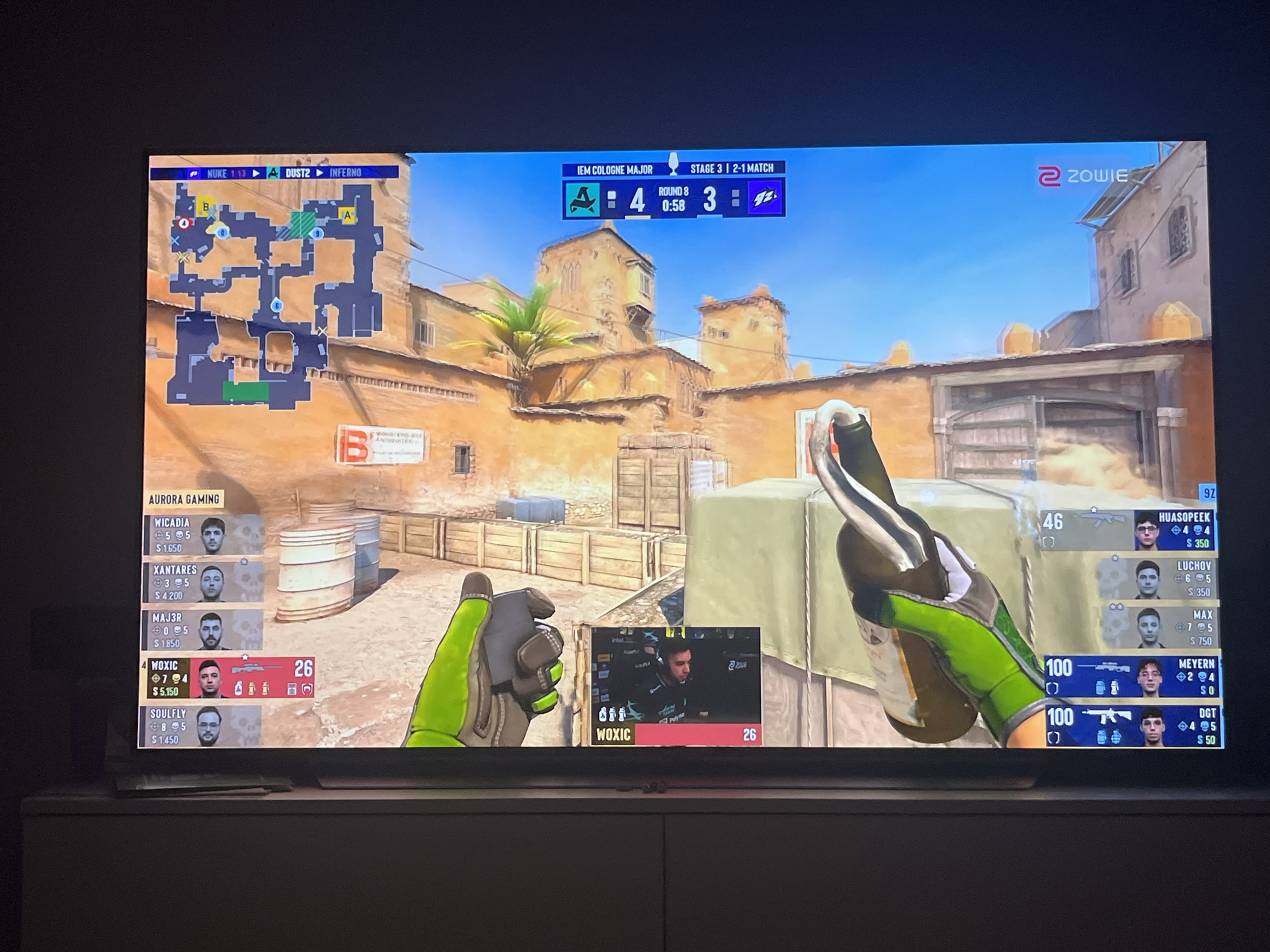 The image shows a television screen displaying a live broadcast of a Counter-Strike: Global Offensive (CS:GO) esports match. The game is being played on the map Inferno. The score is 4-3 in favor of the team represented by the red icon, with the team represented by the blue icon trailing. The match is part of the IEM Cologne Major, Stage 3. The in-game display shows player statistics, including kills, deaths, and money. One player, "Woxic," is highlighted with a kill count of 7 and 3 deaths. A small inset window shows a live stream of "Woxic" playing the game. The screen also displays team names, "Aurora Gaming" for one team. The ZOWIE logo is visible in the upper right corner. The scene is viewed from a slightly elevated angle, suggesting the television is placed on a piece of furniture. The room appears to be dimly lit, with the primary light source coming from the television screen itself. There is no visible information in the image to suggest a connection to Varna, Bulgaria.