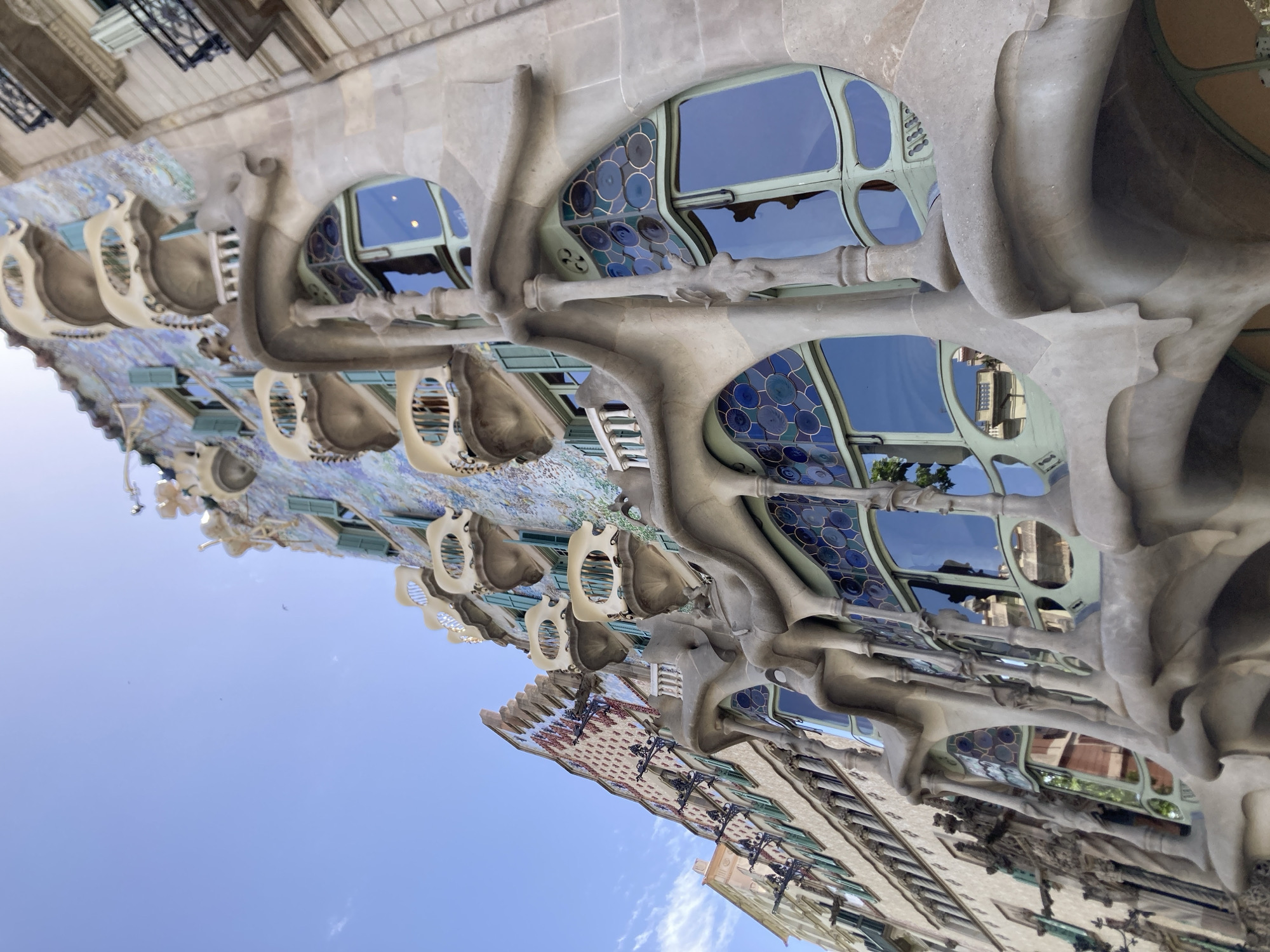 The image shows an exterior view of Casa Batlló in Barcelona, Spain. The architecture is a prime example of Antoni Gaudí's modernist style, characterized by organic, flowing lines and vibrant colors. The facade features a mosaic of iridescent tiles in blues and greens, with sculpted balconies that resemble bones or masks. The windows are large and arched, with intricate stained-glass patterns in shades of blue and green, reflecting the sky.
The photograph is taken from a low angle, looking up at the building against a clear blue sky with a few wispy clouds. The bright sunlight suggests it is daytime, likely late morning or early afternoon. The overall impression is one of wonder and artistry, highlighting the unique and imaginative design of the building. No people are visible in the image, and no text is present. The building's unusual and whimsical nature evokes a sense of fantasy and playfulness.