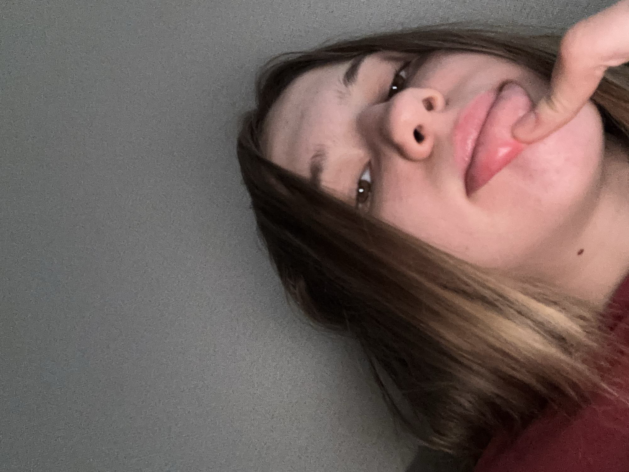 The image is a close-up, low-angle selfie of a young person with medium-length brown hair, looking upwards towards the camera. The background is a plain, textured gray surface, likely a wall or ceiling, and does not provide any specific environmental context. The person's face is partially visible, with their eyes looking towards the lens. Their mouth is open, with a finger touching their lower lip, and their tongue is slightly visible. The lighting appears dim and artificial. The image does not contain any information that can be used to determine its location in Vsevolozhsk, Russia.
