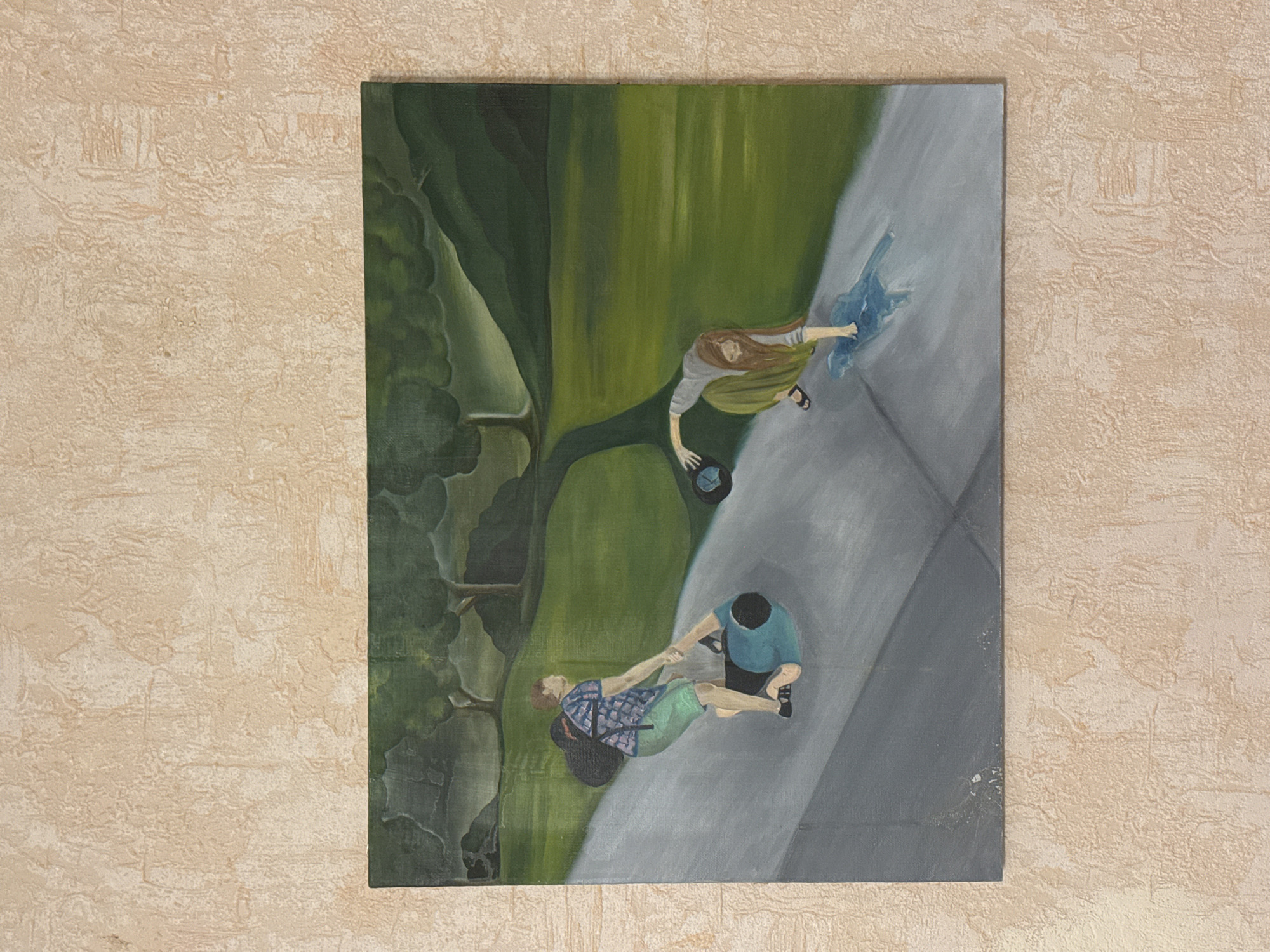 The image is a painting depicting two figures in a park-like setting. On the left, a person wearing a plaid shirt, green shorts, and a backpack is helping another person who is lying on their back in a teal shirt and dark pants. The figures are on a gray paved surface that appears to be a walkway or path. In the middle ground, a path winds through a green grassy area with trees in the background. On the right side of the painting, a person with long brown hair, wearing a gray shirt and a long green skirt, is reaching down towards the paved surface, holding a dark circular object, possibly a hat, in their left hand. There appears to be a puddle or wet spot on the ground in front of this figure. The background shows rolling green hills and trees under a muted sky. The painting is hung on a textured, light-colored wall. There is no indication that this scene is located in the city of Ryazan, Russia.
