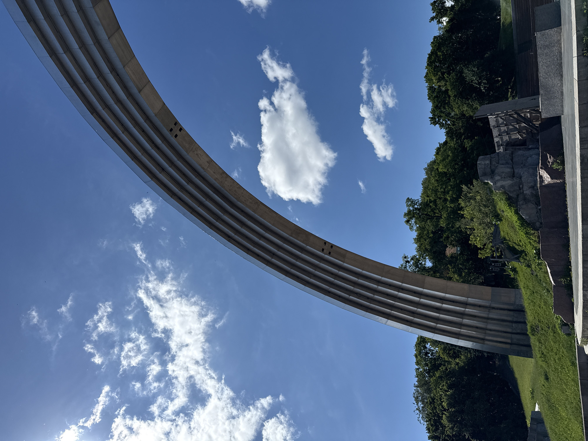 The image shows a low-angle, close-up view of the arch of the Friendship of Nations monument in Kyiv, Ukraine. The massive stainless steel structure dominates the upper right portion of the frame, curving upwards against a vibrant blue sky filled with scattered white clouds. The arch is composed of multiple parallel bands, giving it a ribbed appearance. Sunlight glints off the polished metal surface, highlighting the curvature.
Below the arch, a lush green hillside slopes downwards. Dense, dark green trees form a thick canopy at the base of the hill and extend into the background. In the foreground, there are stone retaining walls and a paved area, suggesting a public space or park. The overall impression is of a monumental structure set within a natural, outdoor environment on a clear, sunny day.