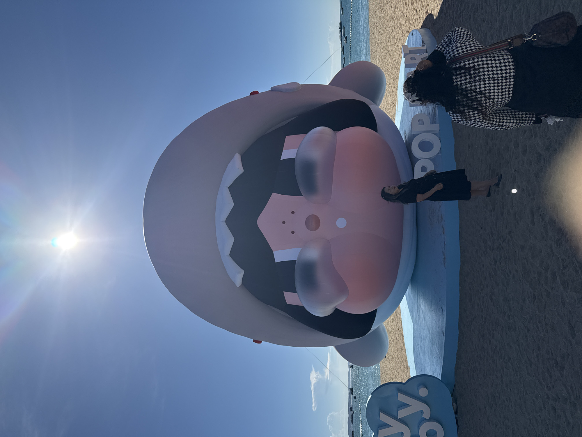An outdoor, daytime, wide shot captures an enormous inflatable cartoon character with its mouth open on a beach in Pattaya City, Thailand. The character is light gray with a large pink lower lip and black interior mouth. The ground beneath the inflatable is a light blue circular platform. To the left of the inflatable, a blue cloud-shaped sign displays the letters "Y Y." To the right of the inflatable, the word "POP ART" is spelled out in white, three-dimensional block letters on the blue platform. The beach sand is light brown. In the background, the blue ocean stretches to the horizon, dotted with several boats. The sky is clear blue with a bright sun causing lens flare at the top center of the frame. Two women are present on the beach, both facing away from the camera. One woman on the left wears a black dress and is positioned closer to the inflatable. The other woman on the right wears a black and white houndstooth jacket and black skirt, with a handbag slung over her shoulder.