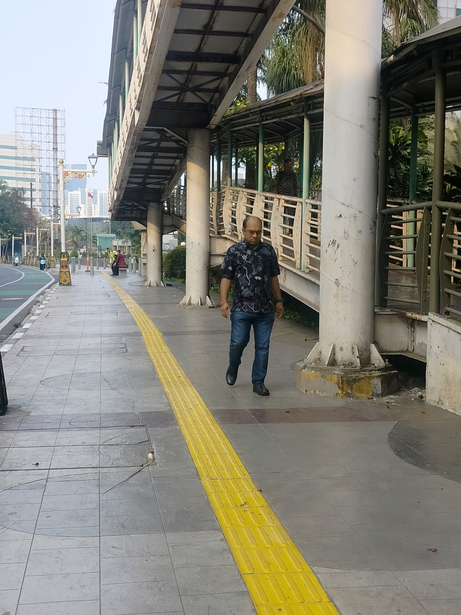 An overhead walkway bridge is the dominant structure in the image, with a man in a dark patterned shirt and blue jeans walking on the sidewalk below. The sidewalk has a prominent yellow tactile paving strip. In the background, there are tall buildings, palm trees, and other pedestrians. The location is identified as South Jakarta, Indonesia.