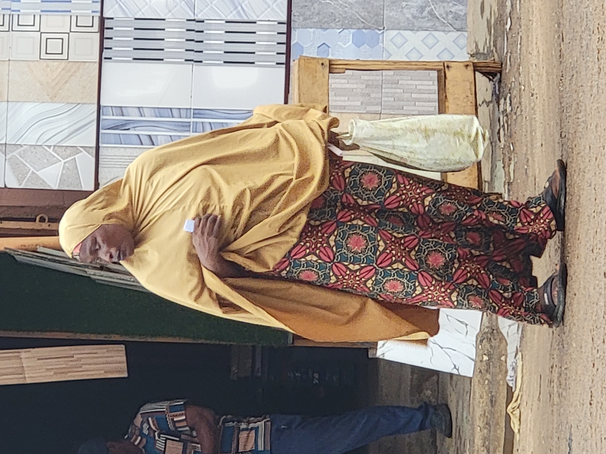 The image, which appears to be rotated 90 degrees clockwise from its intended upright orientation, depicts a scene in Maiburiji, Nigeria, likely at a building materials stall or market.
The primary subject is a woman standing on the right side of the frame. She wears a full, loose-fitting mustard yellow garment that covers her head and body, and a vibrant patterned long skirt featuring maroon, red, green, blue, and gold geometric and floral designs. Her dark-skinned face is visible, and she looks downwards with a neutral or contemplative expression. She holds a light-colored, possibly rolled-up fabric or bag, in her left hand, which rests near her waist. She is wearing dark sandals or flat shoes.
Behind her, and forming the background, is a prominent display of various ceramic tiles. These tiles showcase a range of patterns and colors, including geometric designs with squares and lines in white, grey, blue, and black, as well as wavy and abstract motifs in shades of beige and light brown. A wooden structure, possibly a pallet or stand, is visible near the woman's right foot, indicating a commercial setting for building supplies.
In the lower left foreground, another person is partially visible from the chest down, wearing a patterned shirt with blue, brown, and white geometric designs, and dark blue pants. The ground beneath them is a light brown, earthy or dusty surface. The overall lighting suggests it is daytime, with soft, ambient light illuminating the scene. No discernible text is visible in the image.
