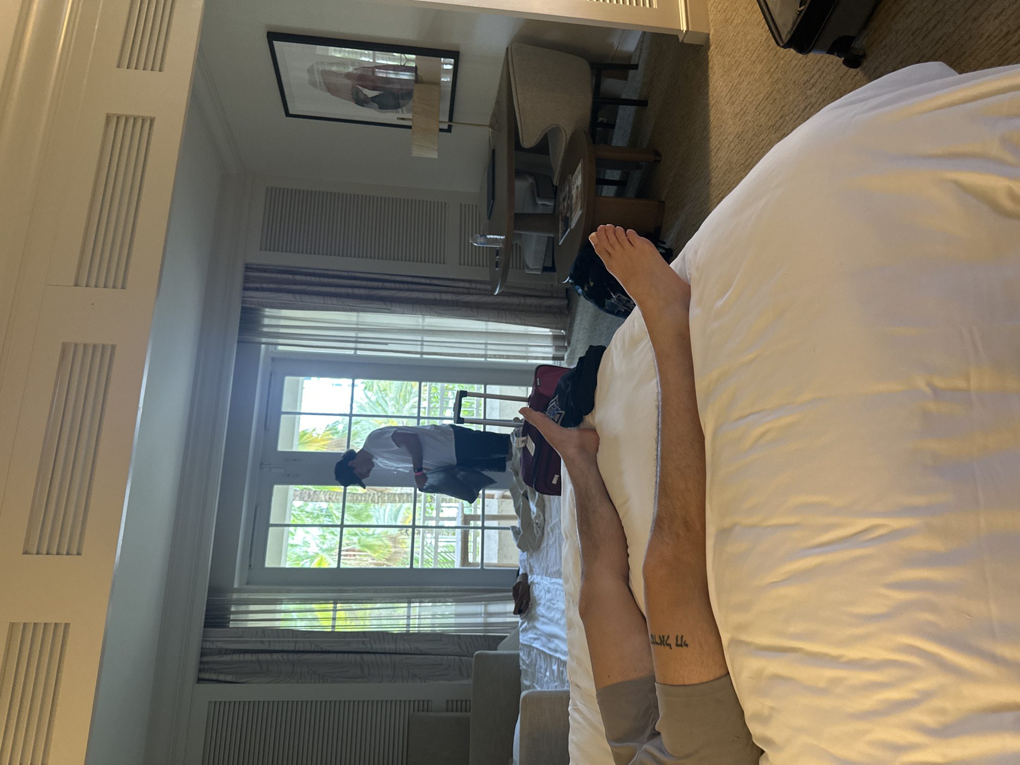 A bedroom scene in Aventura, USA. A person is lying on a bed. The legs of a man are visible, with a tattoo on one leg. Another man is outside the glass doors, partially visible. Luggage is visible near the open doors. The room has white walls and curtains, a desk, and a chair. A framed picture hangs on the wall.