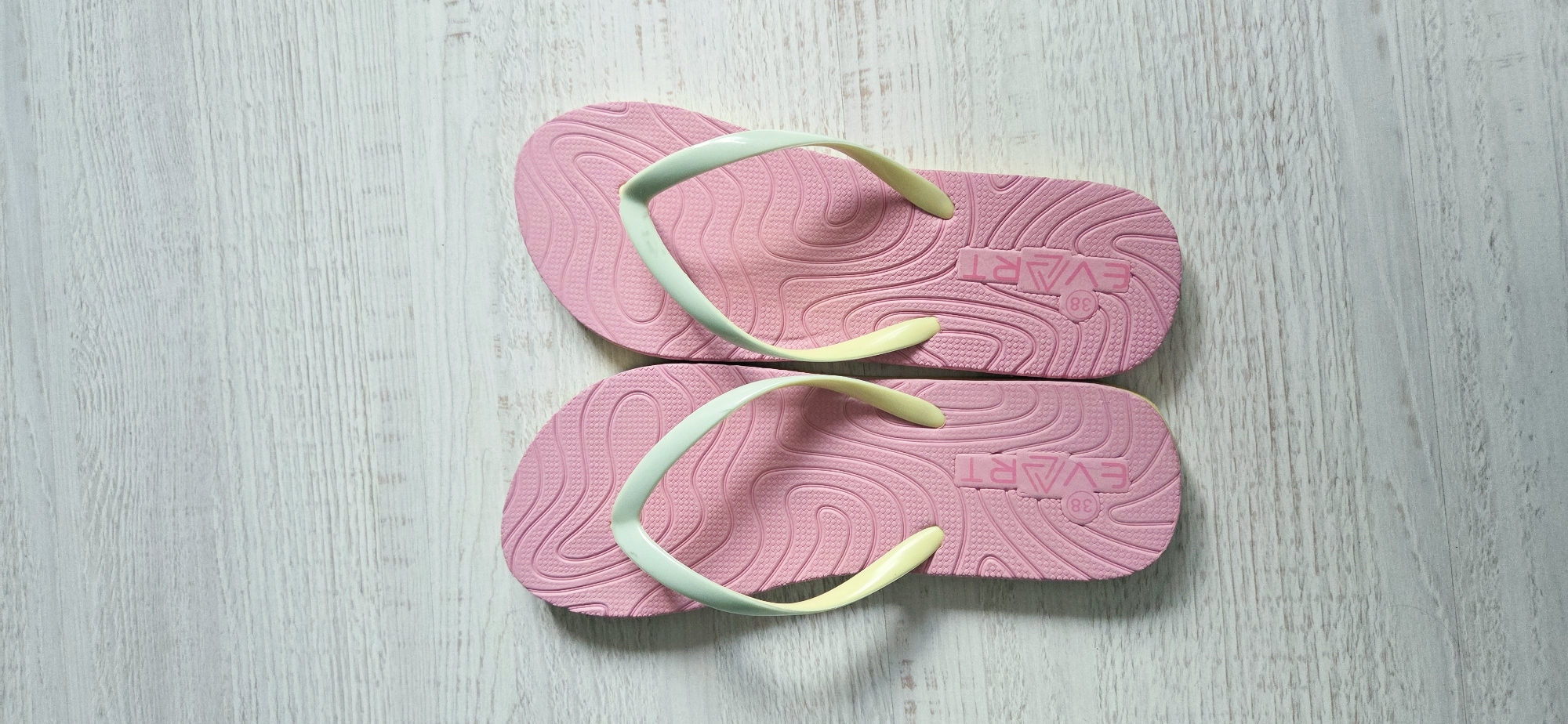 Two pink flip-flops are laid out on a light-colored, horizontally-grained wooden surface. The flip-flops are positioned side-by-side, with the toe of each pointing upwards. Both flip-flops have a light mint green strap that transitions to yellow at the tips, and the soles are embossed with a wavy, textured pattern and the text "EVART" along with the number "38" within a circle. The background is a rustic wooden plank surface, likely a floor or table, with visible wood grain and color variations. The lighting appears to be natural, casting soft shadows. There is no indication of the city of Lipetsk, Russia in the image itself.