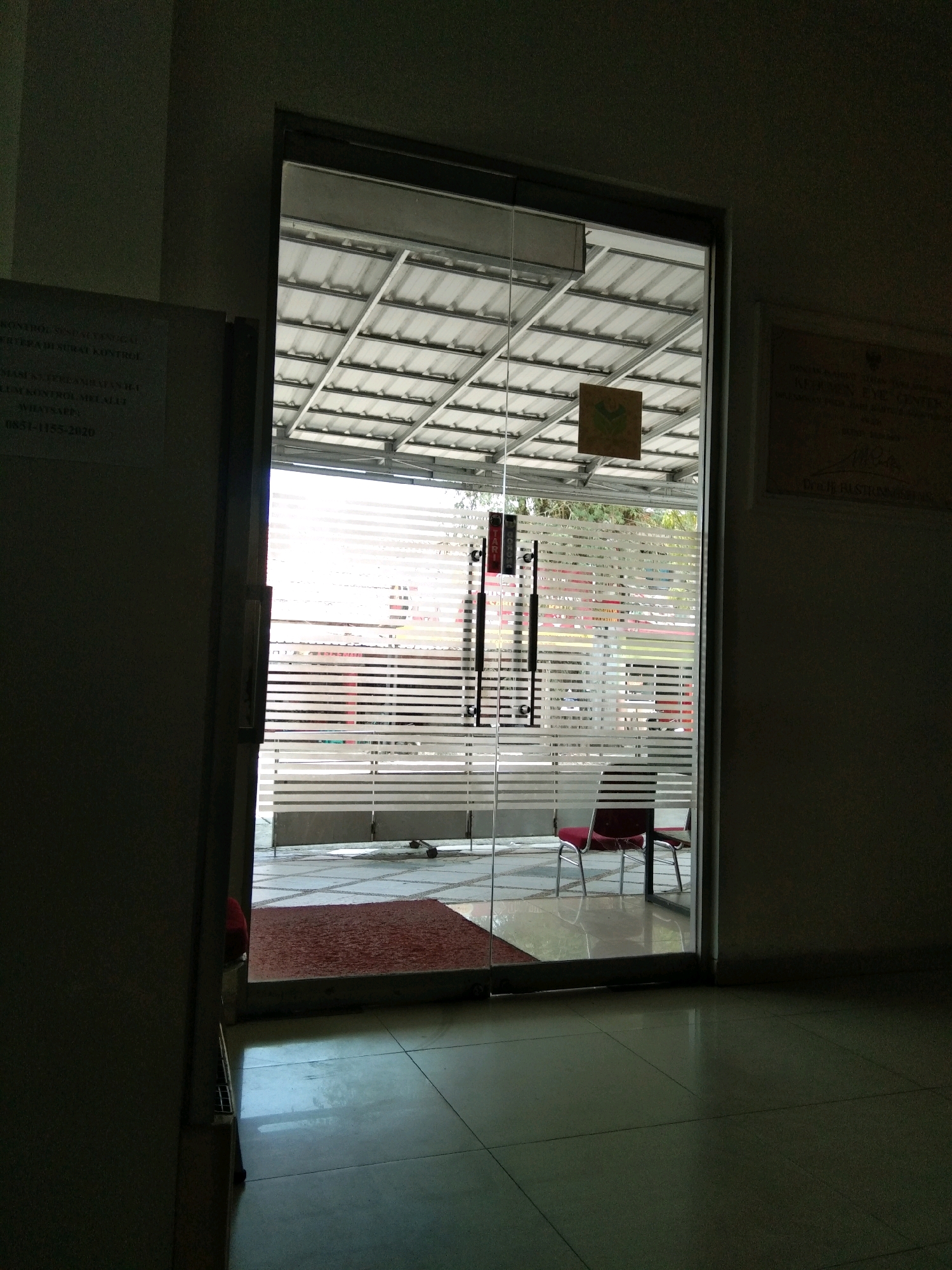 The image shows a dark hallway leading to a glass door with horizontal frosted stripes. Outside the door, there is a covered outdoor area with a metal roof structure. Two red chairs with metal frames are visible, along with a red mat on the tiled floor. A sign with a green logo is affixed to the glass door. On the left wall, a sign with Indonesian text is visible. The floor is tiled with large square tiles. The overall lighting suggests an indoor setting transitioning to an outdoor area with ample natural light. The location is stated to be in the city of Kebumen, Indonesia.