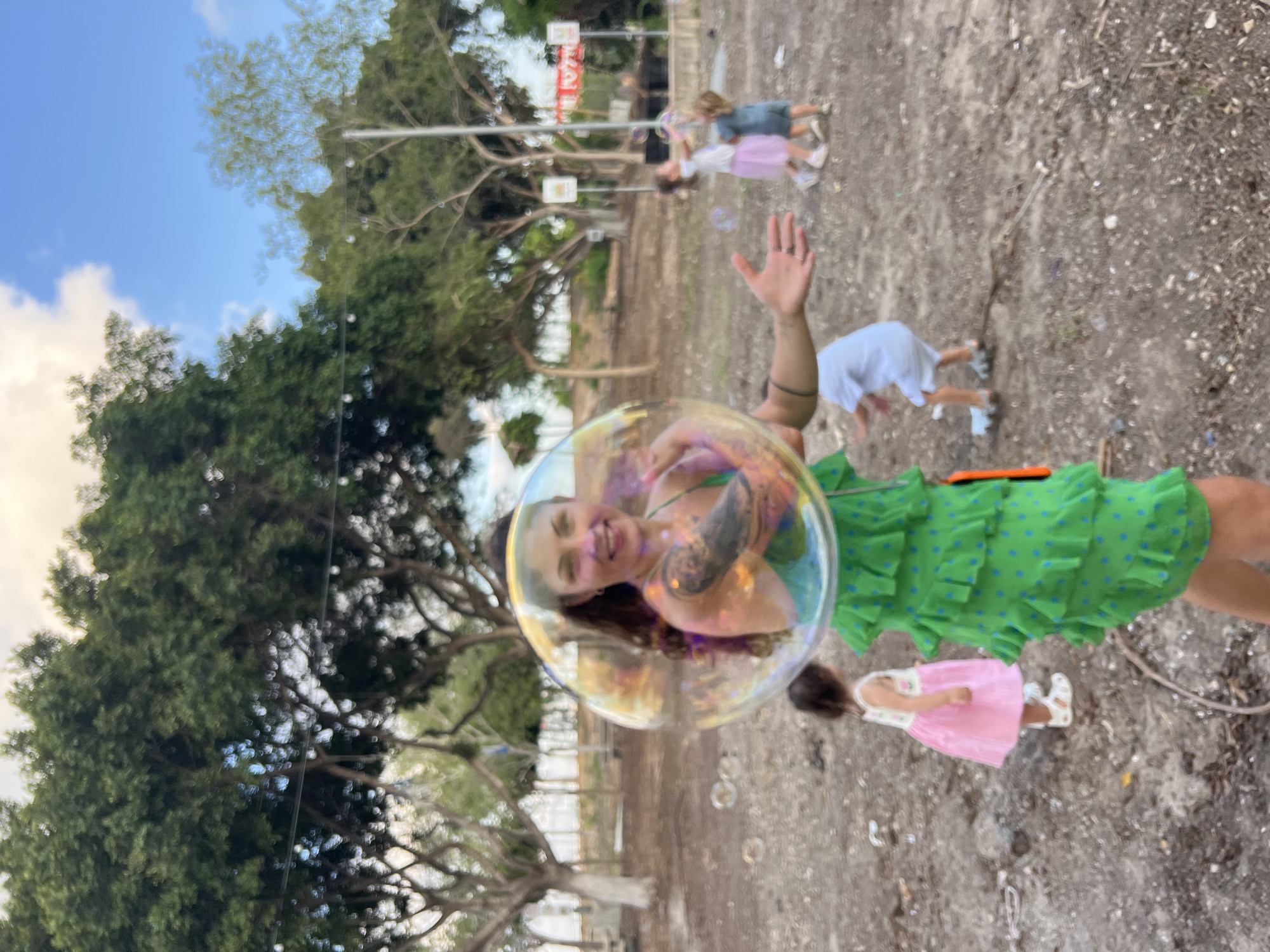 A person is blowing bubbles outdoors in Kiryat Bialik, Israel. The subject, a woman with tattoos, is partially visible within a large, iridescent bubble. She is wearing a green dress with blue polka dots and ruffles. Her right hand is raised, and she appears to be smiling. Several smaller bubbles are scattered in the air around her, and another child in a pink dress is visible in the foreground. In the background, trees and a cloudy sky suggest it is daytime. The ground appears to be a mix of dirt and gravel. A few people and what appear to be signs are visible in the distance.