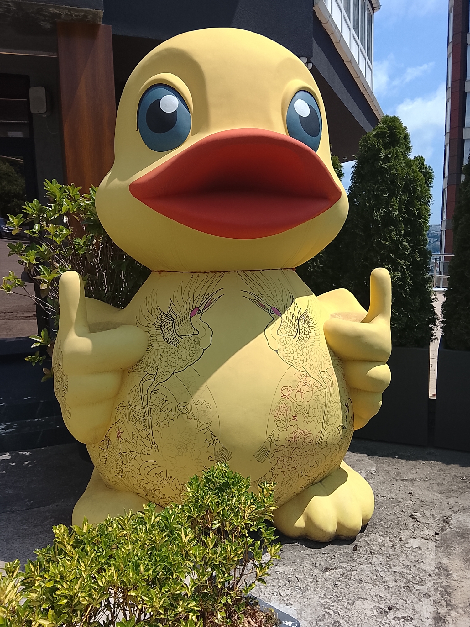A large, bright yellow duck statue is positioned outdoors in front of a building. The duck has prominent blue eyes with black pupils and a large red beak. Its body is decorated with intricate black line drawings of cranes and floral motifs. The statue is giving a "thumbs up" with both of its hands, which are positioned at chest level. In front of the statue, there is a low-lying green shrub. Behind the duck, a multi-story building with white window frames is visible, along with several tall, dark green coniferous trees planted in dark rectangular planters. The ground appears to be paved with concrete. The sky is blue with scattered white clouds. This image was captured in Oryol-Izumrud, Russia.