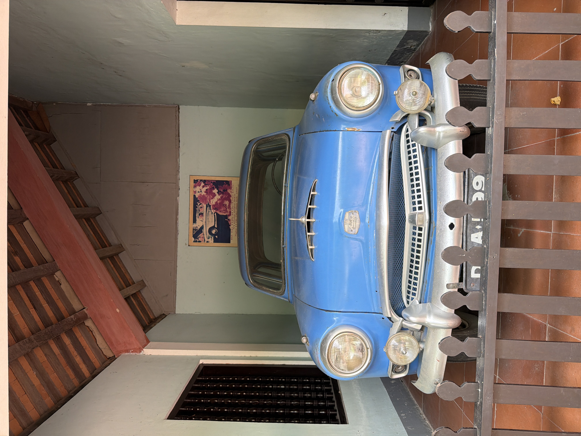 This image shows a vintage blue car parked under a covered structure with a tiled roof in Kim Long Ward, Vietnam. The car is a Kaiser Virginian, recognizable by its distinct grille and dual headlights. It is positioned centrally within the frame, facing forward. The car is protected by a dark brown wooden fence in the foreground. Behind the car, on the wall, is a framed picture of a vintage car and people. The environment appears to be an outdoor exhibit or a historical site, with terracotta tiles on the floor and pale green walls. A wooden lattice window is visible on the left side of the image. The scene is static, with no people or discernible action occurring.