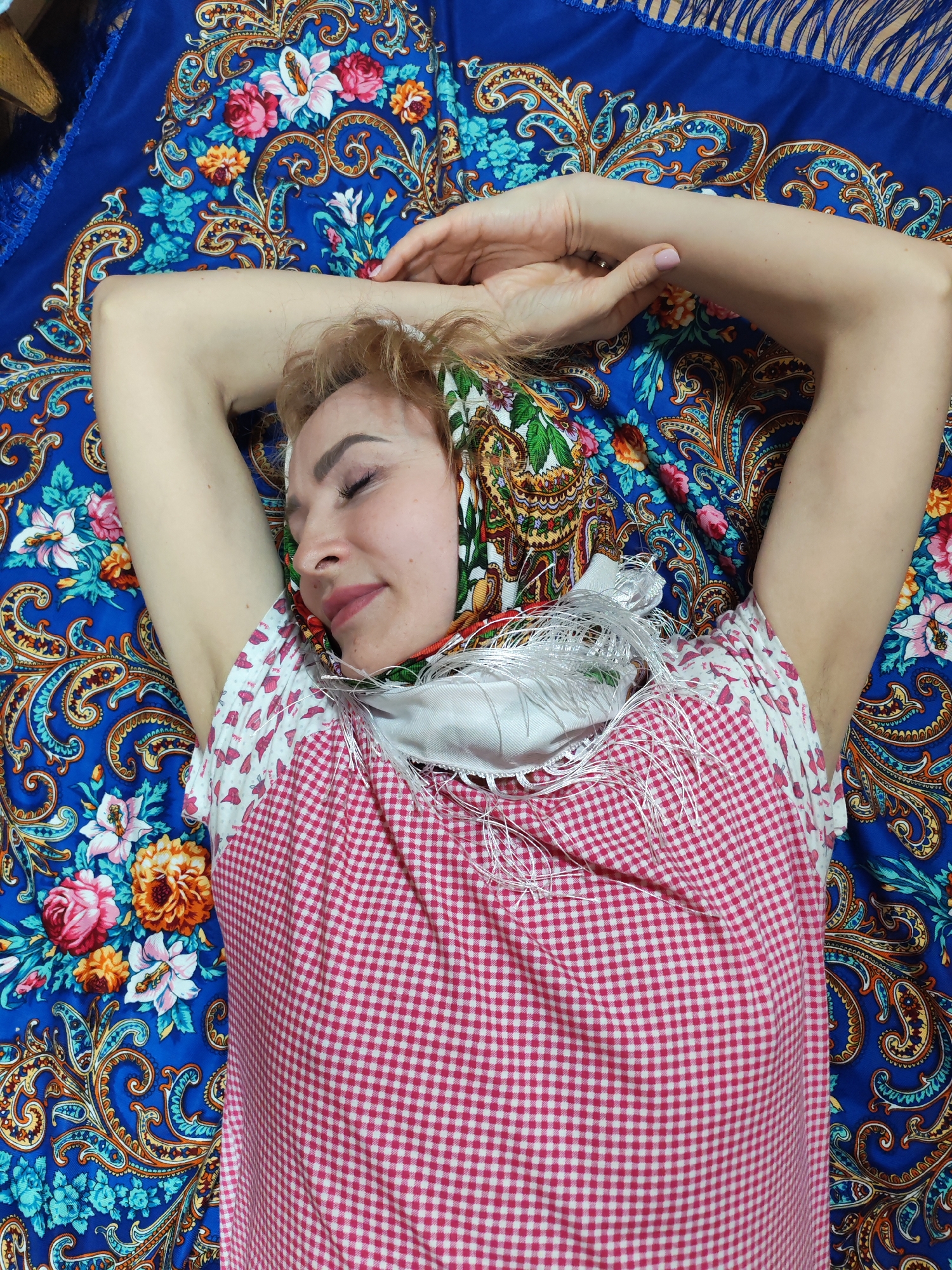 A fair-skinned woman with blonde hair is lying on a patterned blue blanket. Her eyes are closed, and she wears a red and white checkered top with a white and floral headscarf. Her arms are raised and crossed above her head. The blanket beneath her is adorned with intricate paisley designs and floral patterns, with a fringed edge visible at the top. The environment is indoors, and the location is unspecified within the city of Chaikovsky, Russia.