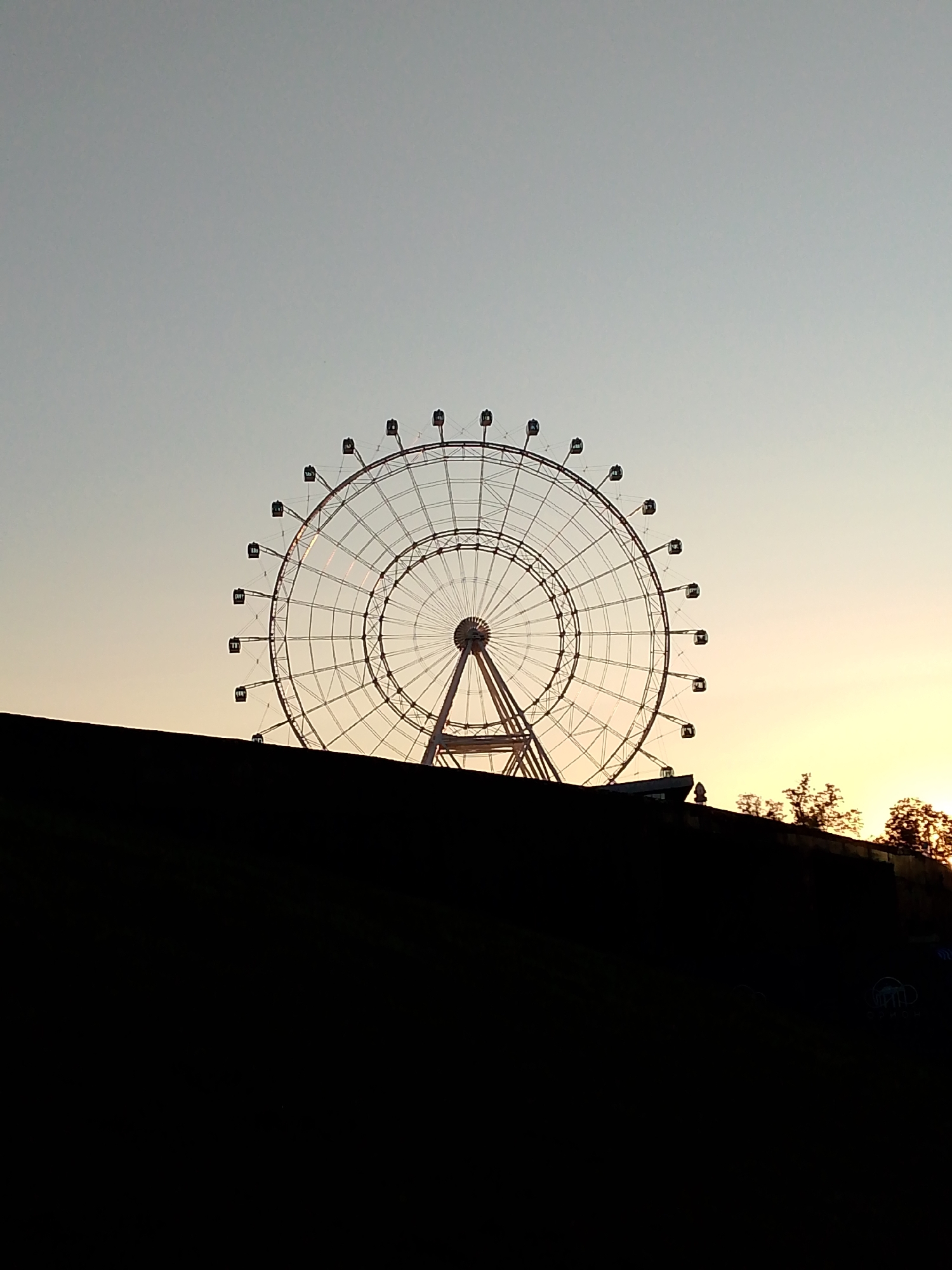 The image shows a ferris wheel silhouetted against a pale sky during what appears to be sunset. The ferris wheel is large and intricate, with many spokes and gondolas. In the foreground, a dark, irregular shape, likely a rooftop or hillside, obscures the bottom portion of the image. Trees are visible on the right side of the image, also silhouetted against the sky. The overall impression is one of a quiet, possibly late evening scene in a city park or amusement area. The specific location in Moscow is not discernible from the image itself.