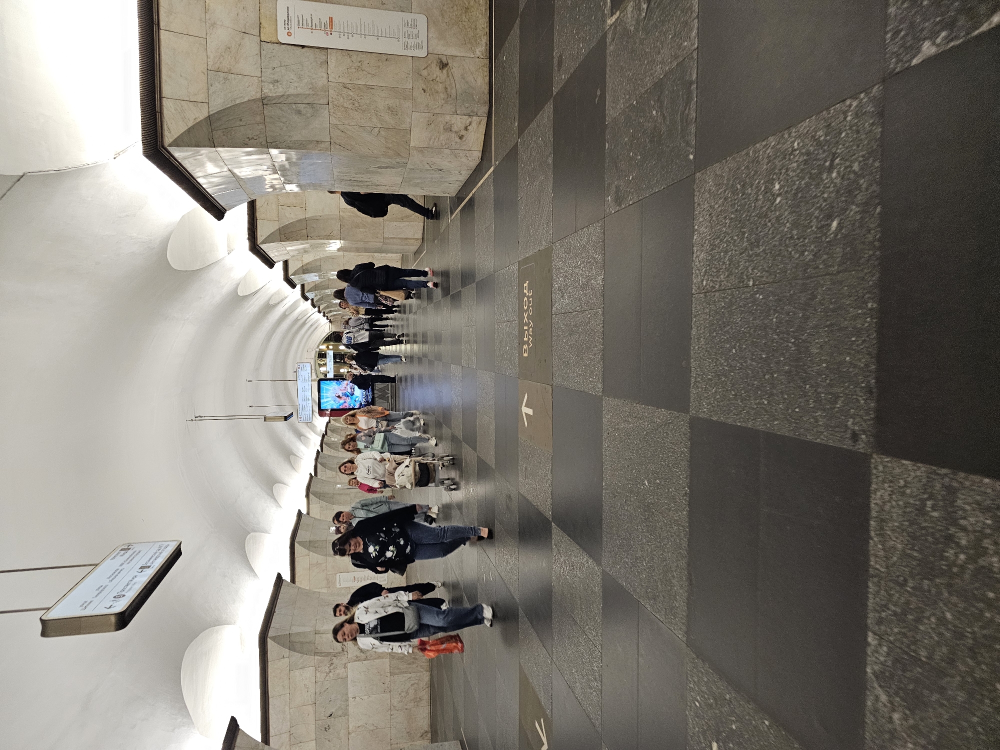 The image depicts the interior of a Moscow Metro station, likely Arbat-Pokrovskaya line, given the distinctive white vaulted ceiling and marble-clad pillars. The station is well-lit, with people walking along the platform. The floor is paved with dark and grey checkered tiles. Several individuals are visible, including a woman pushing a stroller, a couple walking arm-in-arm, and other pedestrians in various directions. Signs are mounted on the walls and ceiling, indicating directions and information relevant to commuters. The architecture is grand, typical of many Moscow Metro stations, featuring arched ceilings and decorative stonework. The overall scene conveys a busy but orderly public transportation environment.