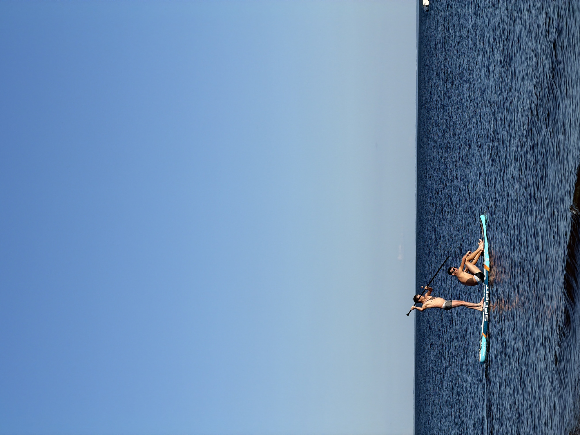 Two men are stand-up paddleboarding on a calm, dark blue body of water under a clear, bright blue sky. The man in the front is seated on the paddleboard, facing the camera and wearing dark swim trunks. The man behind him is standing and holding a paddle, also facing the camera and wearing dark swim trunks. The paddleboard is turquoise and blue with the word "SPORTIT" printed on the side. The horizon is visible in the distance as a thin line separating the water and the sky. The water is slightly rippled.