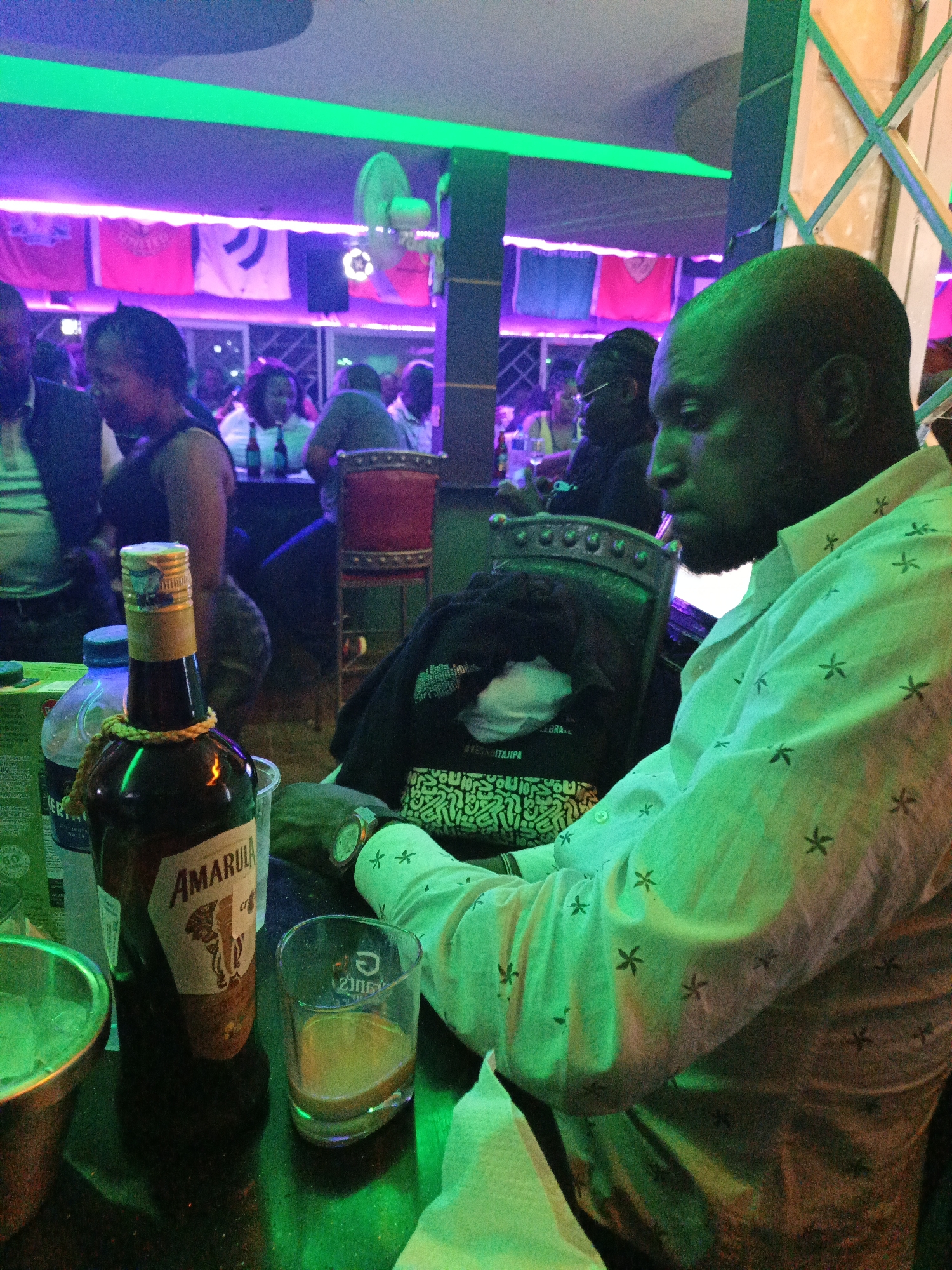 The scene is set inside a dimly lit bar or nightclub in Nairobi, Kenya, characterized by vibrant neon green and purple lighting. In the foreground, a dark-skinned adult male with a shaved head and beard, wearing a light-colored, long-sleeved shirt with small dark star patterns, is seated at a dark table, looking downwards. He wears a metallic wristwatch on his left wrist. On the table directly in front of him are an uncapped bottle of Amarula cream liqueur with a gold cap and a brown rope tied around its neck, a clear glass containing a small amount of light-brown liquid, a clear plastic water bottle, a green carton, and a metallic ice bucket. A crumpled white napkin is also present. A dark garment, possibly a jacket, with visible white text underneath, is draped over the back of his chair. In the midground and background, multiple other patrons are visible, seated at tables in various positions, enjoying the establishment's atmosphere. A woman with dark hair is partially visible to the left of the foreground subject. On a back wall, several flags or banners, illuminated by purple light, are displayed, one of which appears to be a red Manchester United flag. A wall-mounted fan is also visible in the background, along with a decorative, possibly wooden, lattice structure on the right side of the frame.
