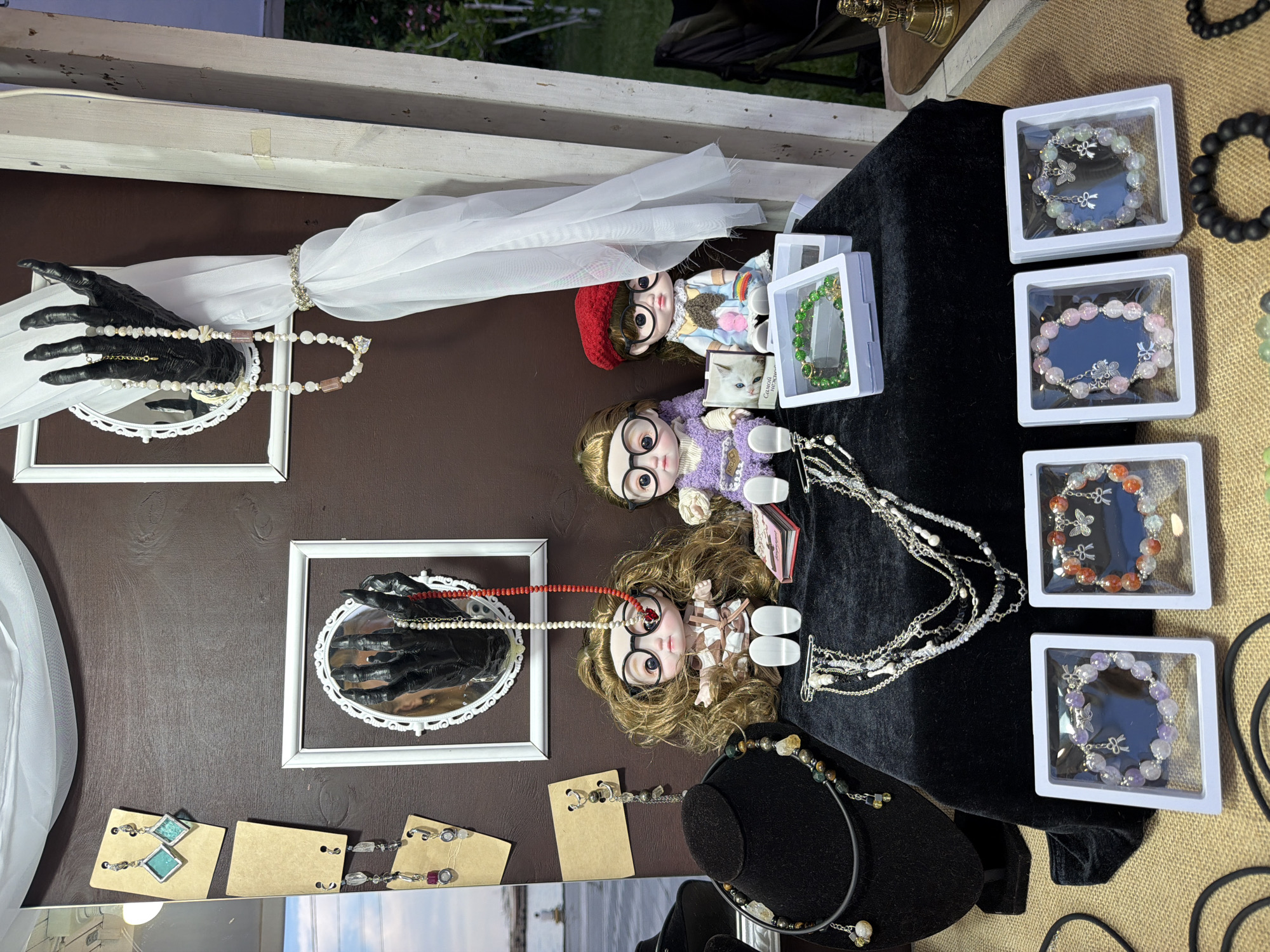The image depicts a display of handmade jewelry, possibly at an outdoor market. The display features two dolls dressed in doll clothing, sitting behind a black velvet cloth. Several necklaces and bracelets are arranged on the cloth and on a black necklace stand. Some of the jewelry is displayed in clear plastic boxes. Behind the dolls, a large black mannequin hand is holding several strands of pearls and beads. On the wall behind the display, there are some earrings hanging on cardboard. The background includes a dark brown wall, a white draped fabric, and a wooden post. The scene appears to be set outdoors, with some greenery visible in the background. There is no indication of the city of Yalta, Ukraine.