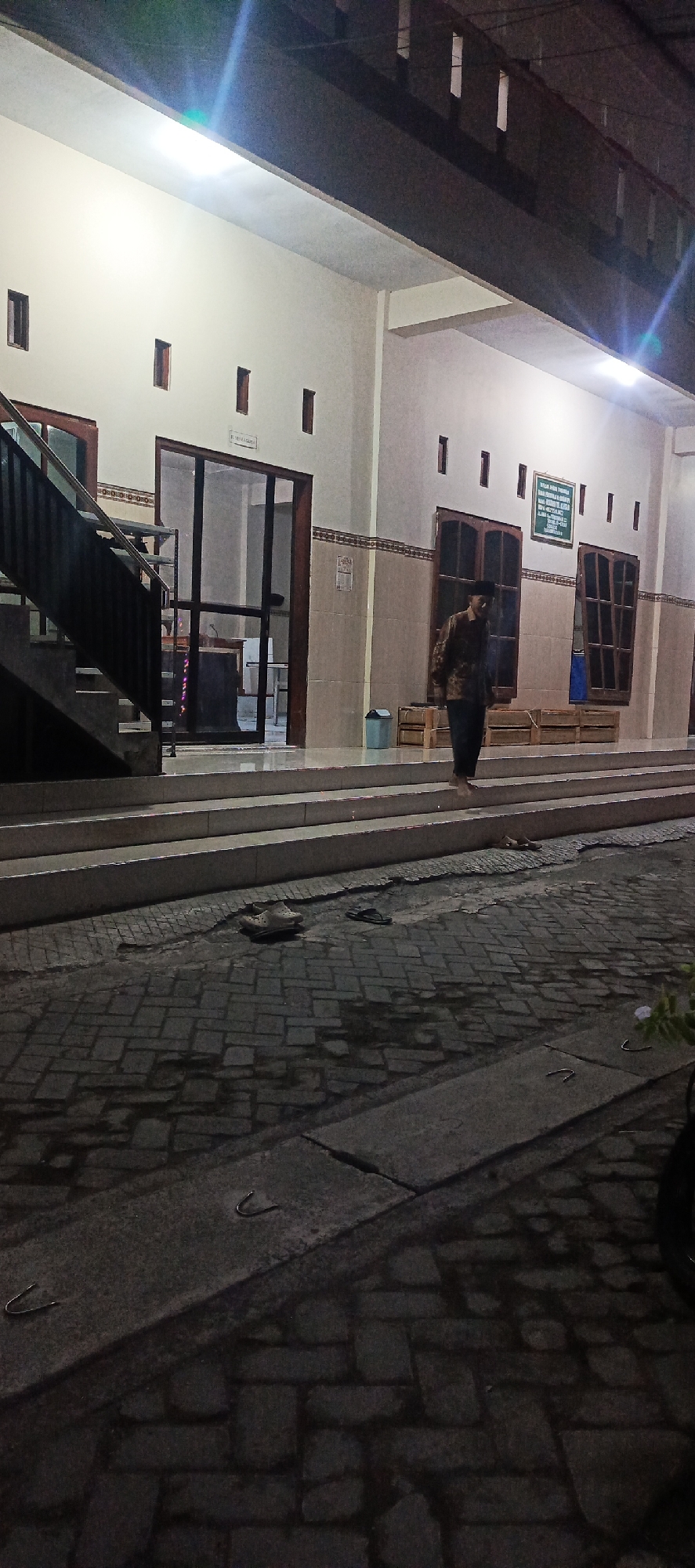 An exterior view of a building, possibly a mosque or community center, at night, in the city of Tempel, Indonesia. A man in a patterned brown and black long-sleeved shirt and dark pants walks down a set of stairs. He is wearing a black skullcap. The building has a light-colored exterior and is illuminated by artificial lights. There are several windows visible, some with wooden frames. A green sign with white text is mounted on the wall. Below the stairs, a paved area with brick-like patterns is visible. Several pairs of footwear, including sandals and clogs, are placed on the ground. A blue trash bin is next to a wooden crate. The scene suggests a quiet evening at a public building.
