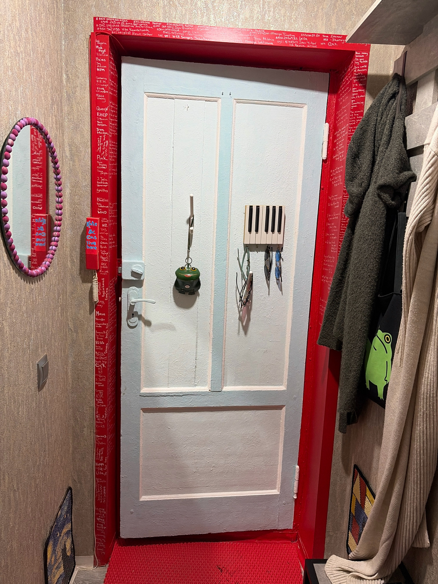 This image depicts a hallway with a red-trimmed door. The door itself is painted white with a light blue frame. On the red trim of the door, various names and phrases are written in white. A pink beaded mirror hangs on the left wall. To the right of the door, several items are hanging on a grey rack, including a dark green sweater and a light beige cardigan, along with a black bag with a green frog design. A small keyholder designed like piano keys is mounted on the door, with several pairs of sunglasses hanging from it. A green frog-shaped keychain is also hanging from a hook on the door. A red doormat is on the floor in front of the door. The walls are textured, and a light switch is visible on the left wall. A small artwork with a mosaic pattern is leaning against the wall on the right. The scene appears to be an interior residential hallway.
