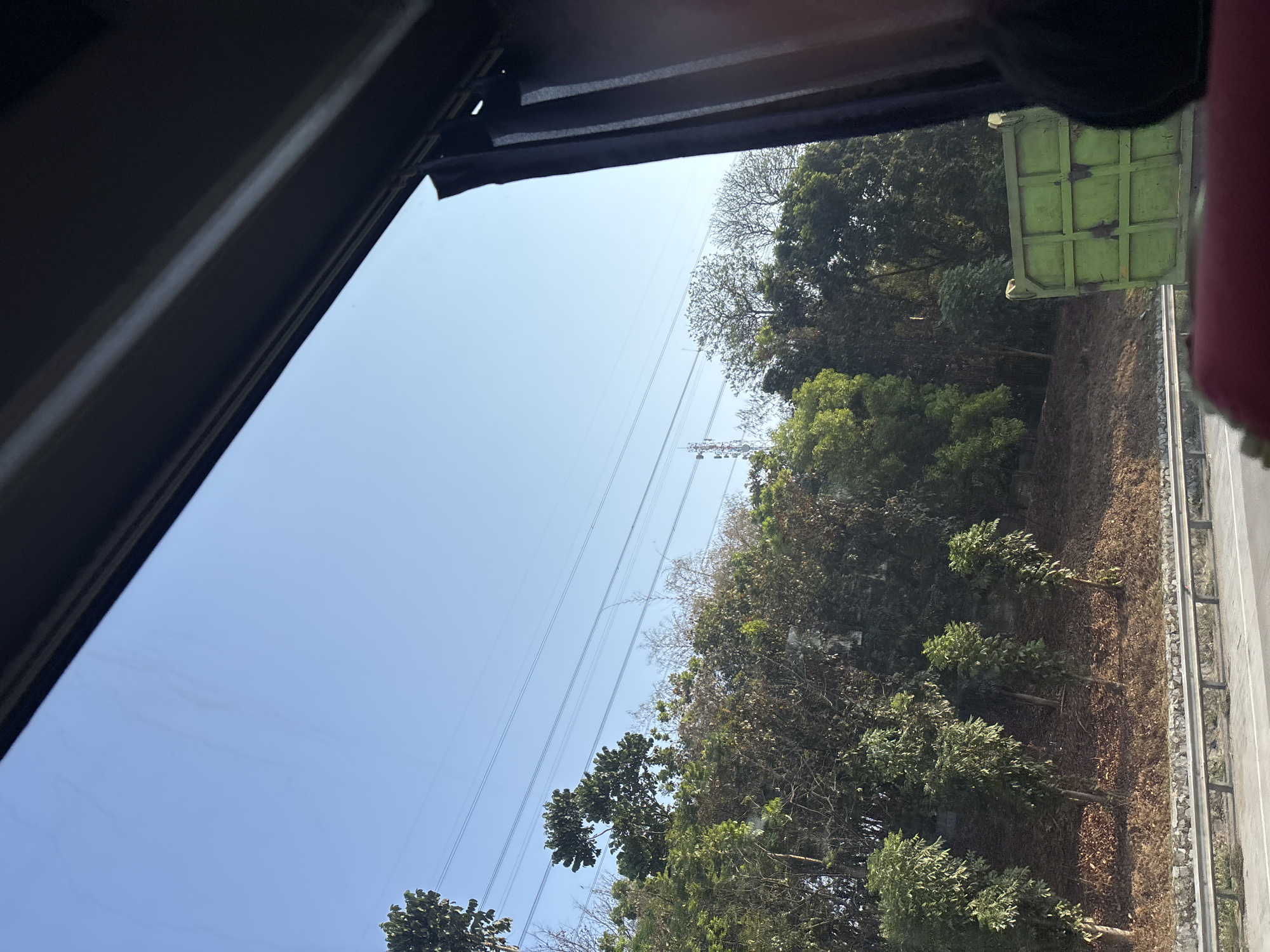 This image shows a view from inside a vehicle looking out at a roadside landscape in Karawang, Indonesia. The foreground is dominated by the interior of the vehicle, with a dark frame on the right and a glimpse of a red seat and a black headrest in the lower right corner. Beyond the vehicle's window, a clear blue sky stretches across the top half of the image.
Below the sky, a dense line of trees and vegetation lines the side of a road. The trees vary in size and density, with some appearing taller and fuller than others. Among the trees, several power lines are visible, strung across the sky and supported by distant poles. A telecommunications tower can be seen in the background, peeking through the trees.
The ground slopes upwards from the road, covered in a layer of dry leaves and interspersed with more trees and bushes. A metal guardrail separates the road from the vegetated embankment. On the right side of the road, a portion of a green dump truck is visible, its large, reinforced back facing the viewer. The scene suggests a rural or semi-rural area along a highway in Karawang, likely during daylight hours due to the bright sky and clear visibility.