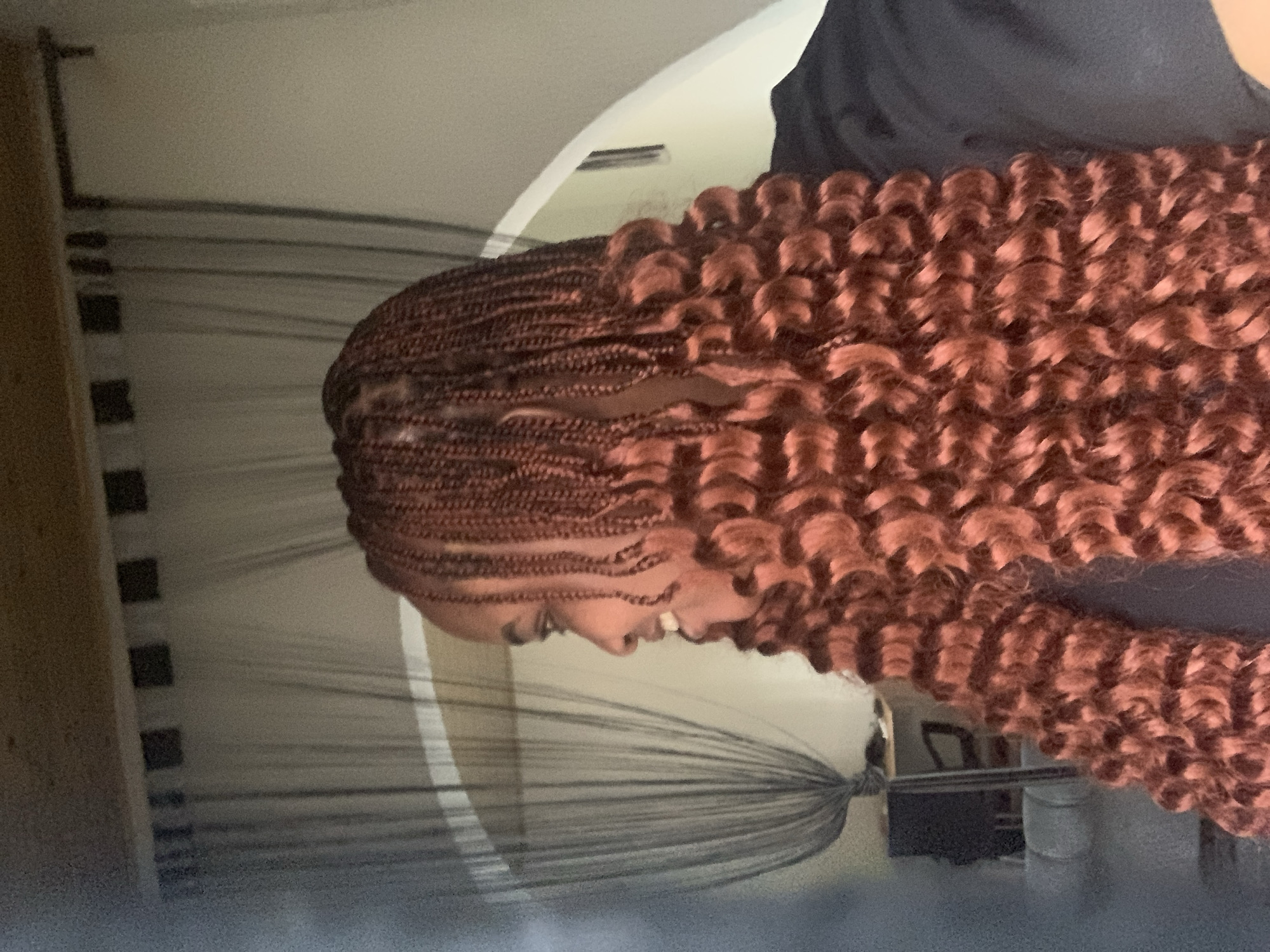 The image shows a person with copper-colored braided hair with curly ends. The hair is styled close to the scalp in neat braids, transitioning into voluminous, curly sections. The person is wearing a dark-colored top. The background consists of a cream-colored wall with horizontal lines. A portion of a light-colored object, possibly furniture, is visible on the left side of the frame. The setting is likely indoors. There is no information provided to confirm if the image was taken in Barakin Akawo, Nigeria.