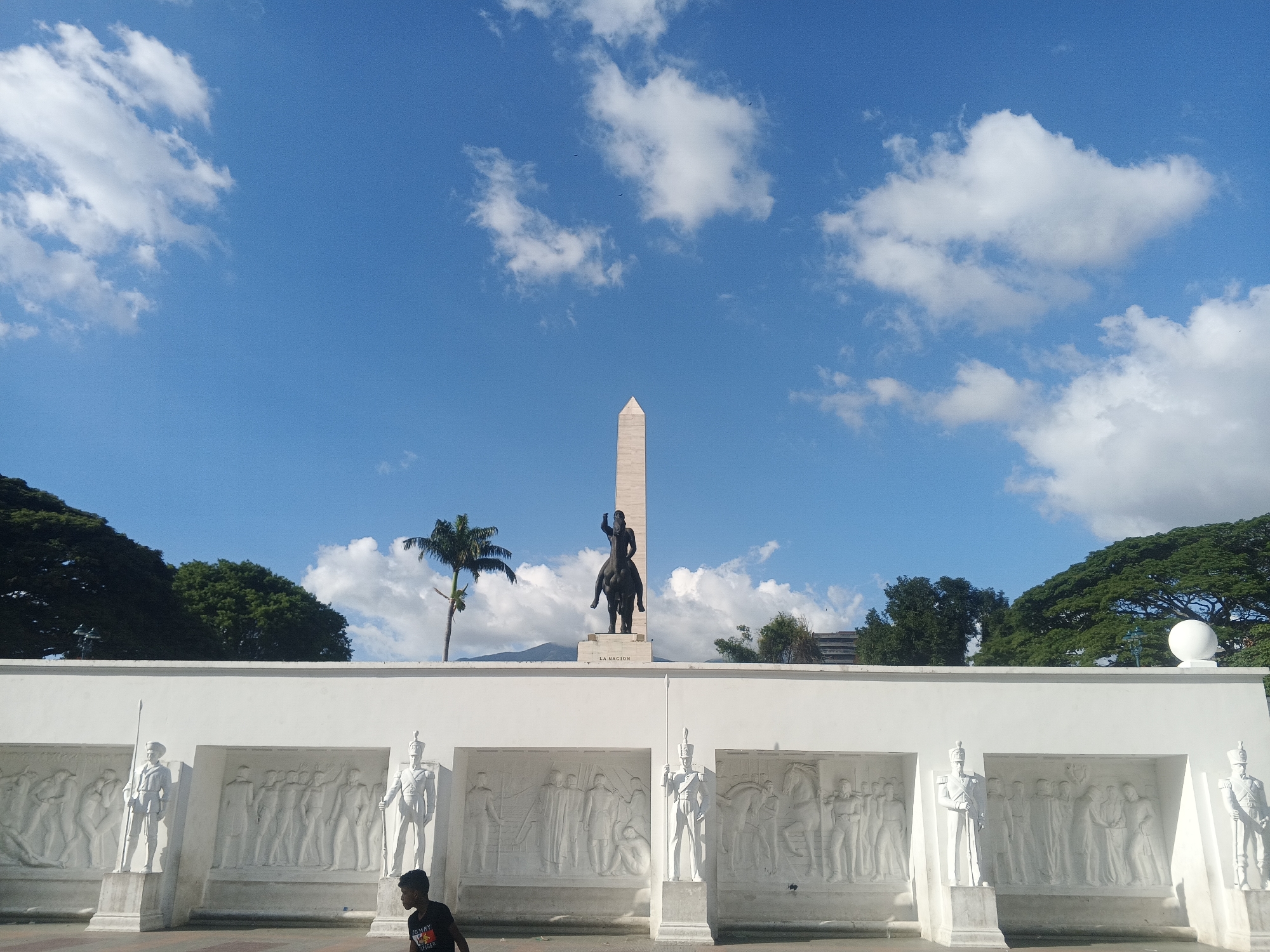 The image shows the Paseo Los Próceres monument in Caracas, Venezuela. The foreground features a white wall with bas-relief carvings depicting historical scenes and figures. White statues of soldiers stand on either side of the wall. In the background, a tall obelisk is visible, with an equestrian statue in front of it. A palm tree and other trees are scattered around the monument. The sky is bright blue with scattered white clouds. A young person wearing a black Tommy Hilfiger t-shirt walks in the lower left corner of the image. The scene appears to be a sunny day in Caracas.