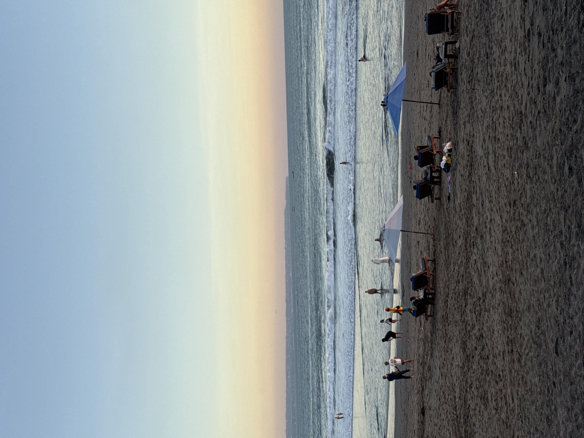 This media file depicts a beach scene in North Kuta, Indonesia. The image is oriented vertically, showcasing a wide expanse of dark sand leading to the ocean. The sky transitions from a pale blue to a soft yellow-orange hue, suggesting either sunrise or sunset. Gentle waves roll onto the shore, with several people visible in the shallow water.
On the beach, a few beach chairs are set up, some occupied by individuals relaxing. A blue umbrella provides shade for one of the seating areas. Scattered across the sand are small groups of people, some standing and others in motion, possibly engaged in activities or conversing. One individual appears to be in a supine position on a chair, while others are walking along the shoreline.
The overall atmosphere appears serene and leisurely, consistent with a beach setting. The lighting and colors suggest a calm, pleasant time of day. There is no visible text in the image. The presence of beach chairs and umbrellas indicates a developed tourist area.