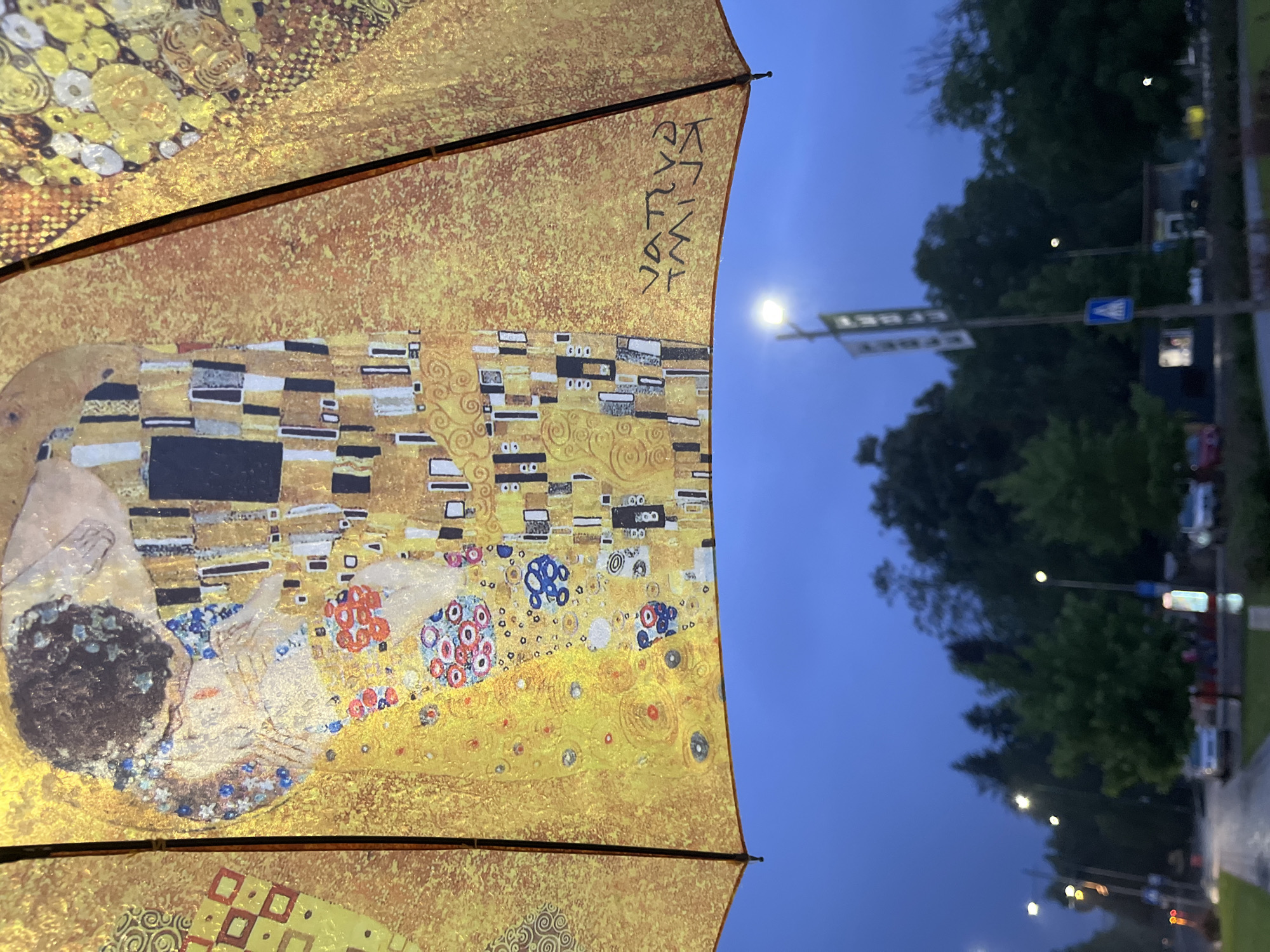 The foreground of the image features an open umbrella, prominently displaying a vibrant, golden-toned artistic print. The print is a reproduction of a section of Gustav Klimt's "The Kiss," showcasing the intricate patterns, golden hues, and decorative elements characteristic of the painting, including circular and rectangular motifs in black, white, blue, and red. Dark ribs of the umbrella are visible, segmenting the artwork. On one golden panel of the umbrella, black stylized text is visible, which appears to read "KATYA JUTYA" or similar names.
The background is an outdoor scene under low light, suggesting dusk or evening. A dark blue sky occupies the upper right portion, with several illuminated streetlights casting a soft glow. Details in the background are blurred, but reveal trees, a road or street, and parked cars along the roadside. Two street signs are discernible: one is a blue square with a white pedestrian crossing symbol, and another, on a dark green background, shows the white letters "JBEET" in the provided image orientation. The presence of the open umbrella implies a rainy or recently rainy atmosphere, though no active precipitation is visible. No people are directly visible in the scene.