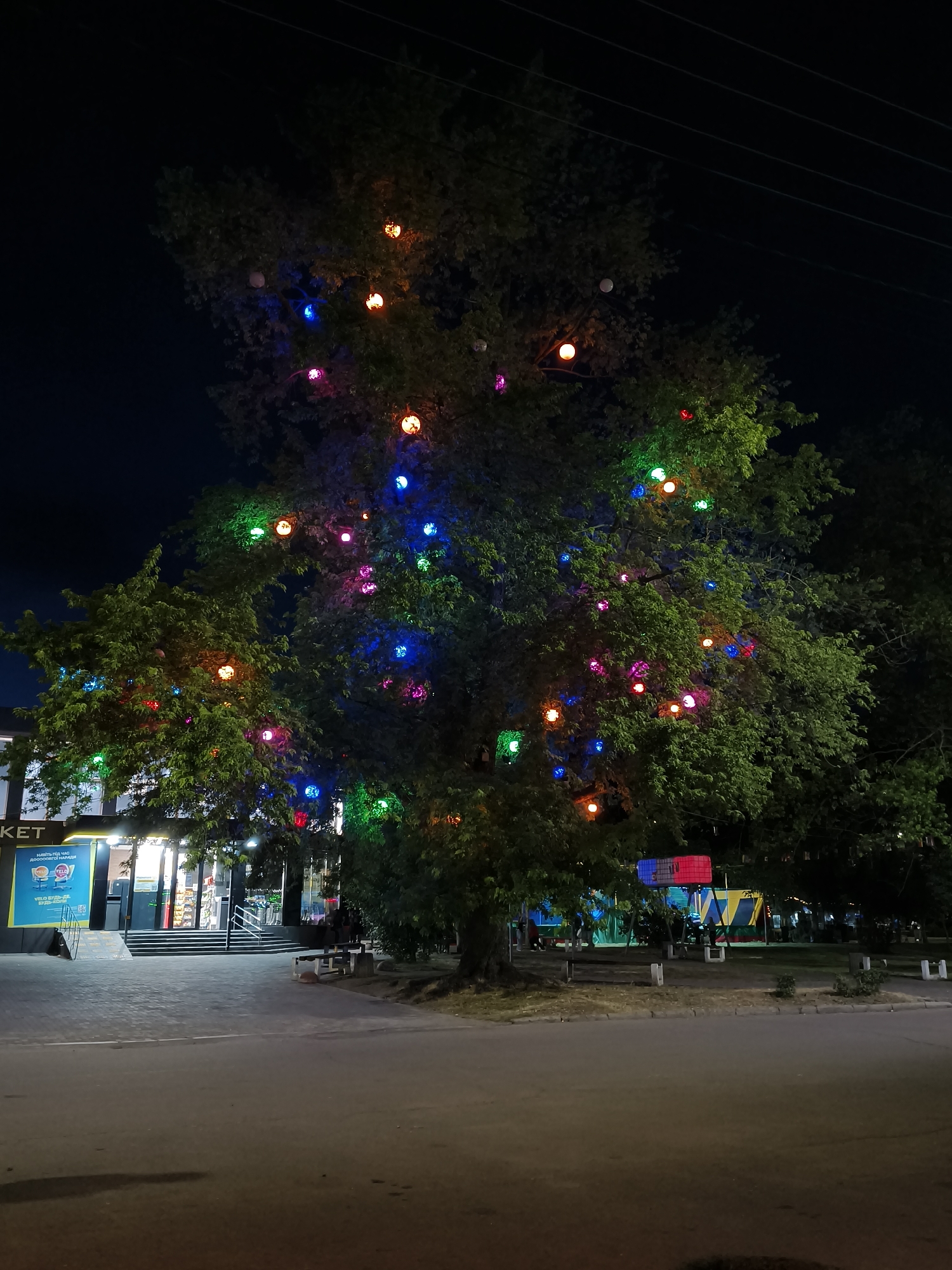An outdoor nighttime scene in Kryvyi Rih, Ukraine, shows a large tree adorned with numerous colorful, spherical lights. The tree is situated in front of a building with a sign that reads "Market." The building appears to be a store or supermarket with illuminated windows and an entrance. To the right of the tree, there is a park-like area with some benches and a colorful, tent-like structure. The ground in the foreground is a paved road. The sky is dark, and power lines are visible above the tree. The lighting from the tree and the building creates a festive and somewhat magical atmosphere. There are a few people visible in the background, appearing to be walking or standing near the market entrance and in the park area. The overall impression is one of a public space decorated for an event or simply for ambiance.