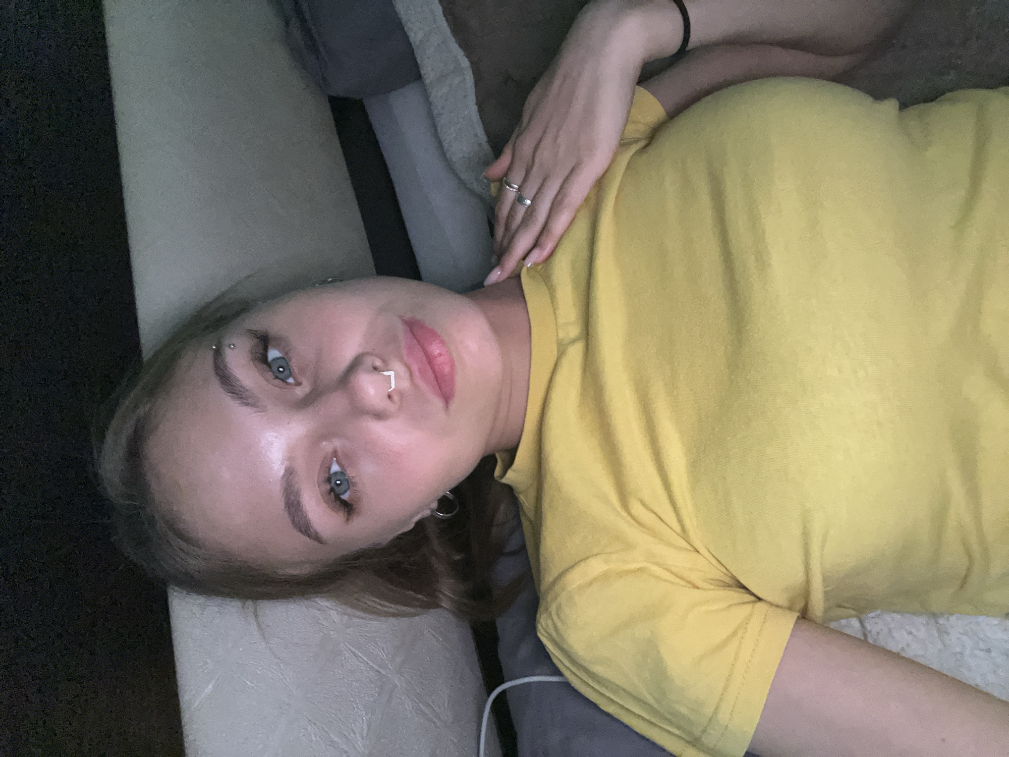 A woman with light skin and blue eyes is lying on a sofa. She is wearing a yellow t-shirt, rings on her fingers, and has an eyebrow piercing and nose ring. The background includes a couch with various pillows and blankets. The location is not discernible from the image, so it is not possible to determine if it is in the city of Фоки, Russia.
