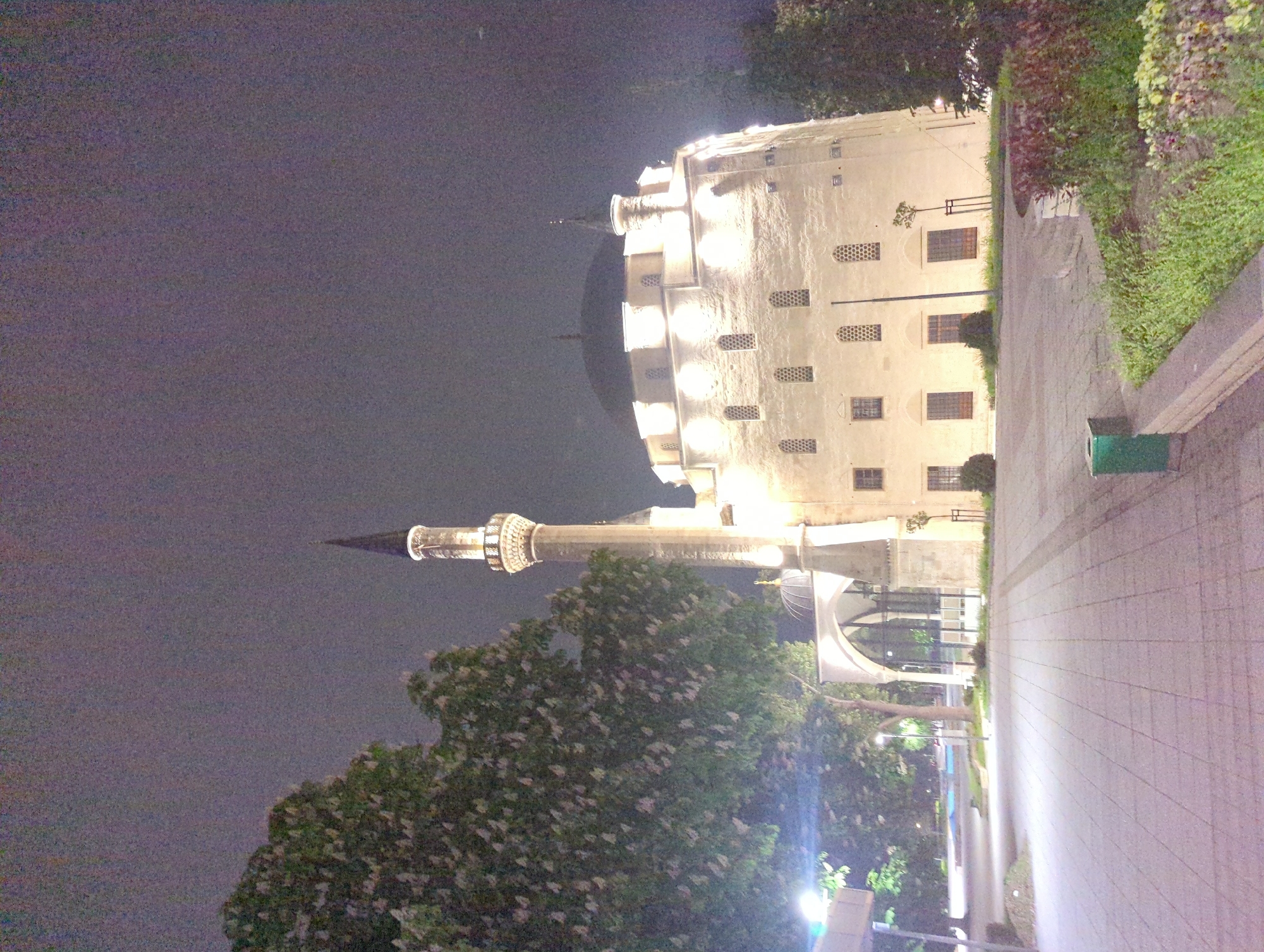 The image depicts the Ibrahim Pasha Mosque in Razgrad, Bulgaria, at night. The mosque's cylindrical main structure and minaret are illuminated, contrasting with the dark sky. A paved walkway runs alongside the building, bordered by greenery. Trees with flowering blossoms are also visible in the foreground.