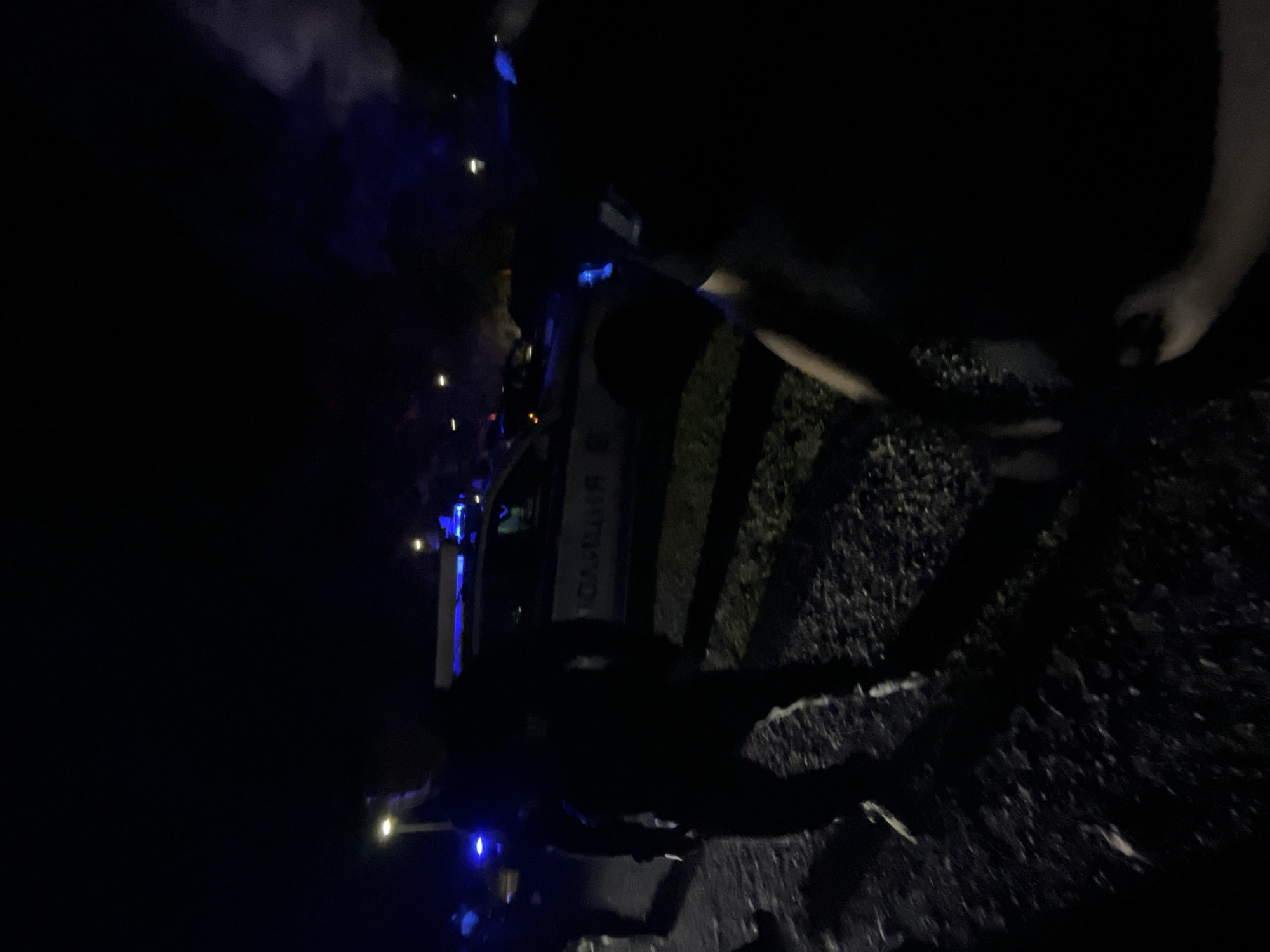 Captured at night, the image depicts an ambulance illuminated with blue lights. The setting appears to be outdoors, possibly a roadside or parking area, with dark ground visible. There are obscured figures surrounding the ambulance and near the back passenger of the vehicle. The text on the side of the vehicle reads “Бърза Помощ.” It is located in Burgas, Bulgaria. The image is underexposed and lacks clear details.