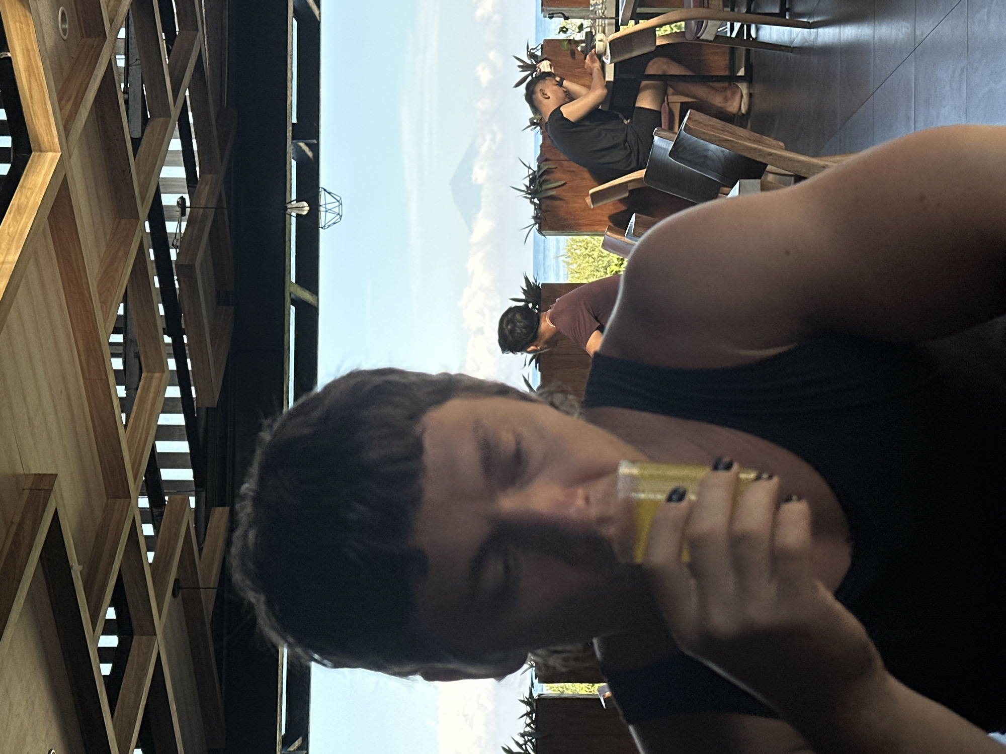 A rotated medium shot depicts a person in a black tank top drinking from a small glass. The subject has dark hair. In the background, other individuals are seated at wooden tables, with a distant view of the ocean and a mountain partially obscured by clouds. The scene appears to be an open-air restaurant with a unique geometric wooden ceiling. The city is Unknown, Indonesia.