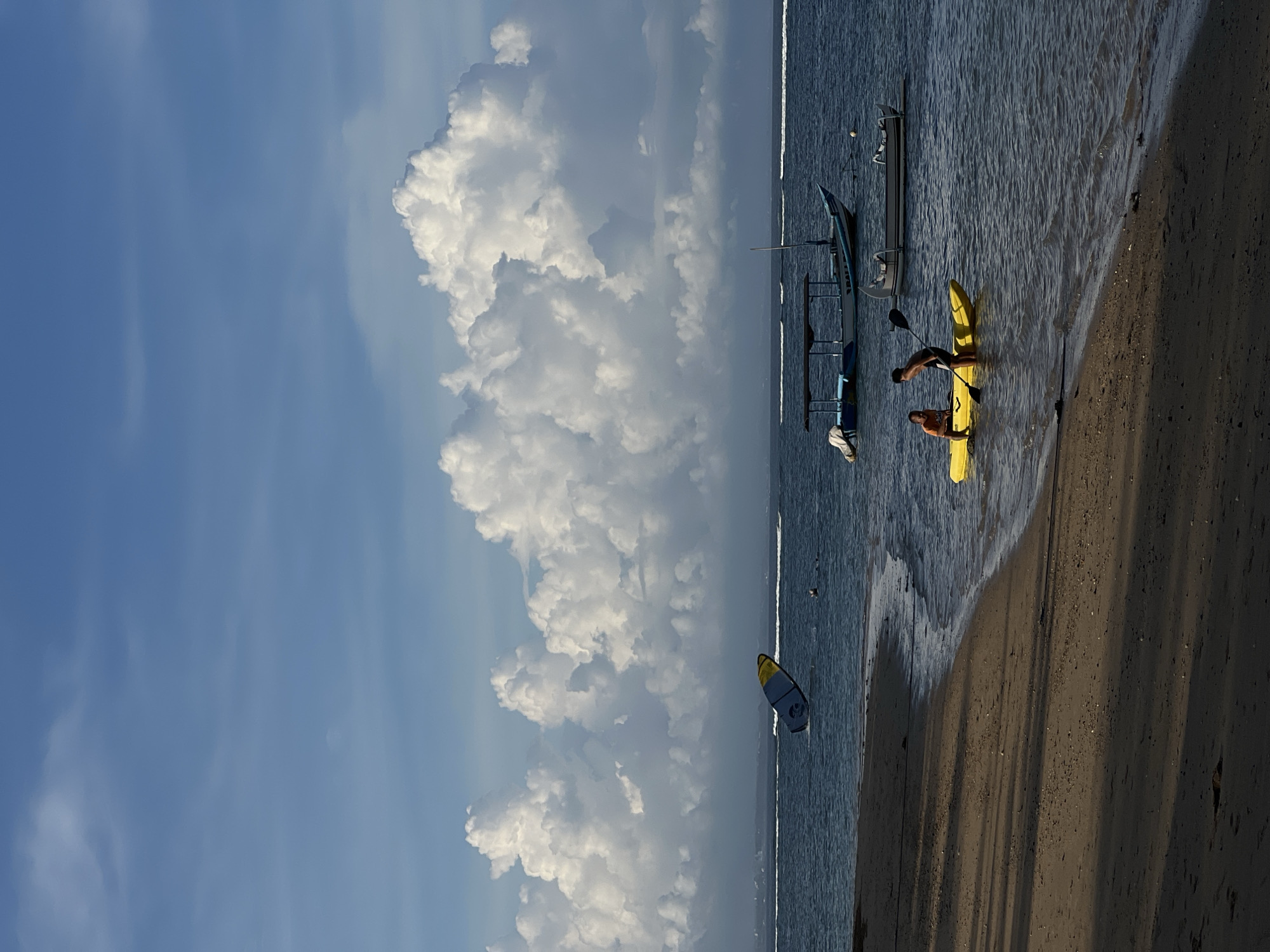 The image shows a beach scene in Denpasar, Indonesia. A large cumulus cloud formation dominates the left portion of the frame, contrasting with a clear blue sky. The right side of the image features the coastline with a dark sand beach and blue ocean. Several boats are visible in the water, including a traditional outrigger canoe and a yellow kayak. Two people are near the kayak, one appearing to be pushing or paddling. A kite-surfing board is floating in the ocean further out. The shoreline has gentle waves lapping at the sand.