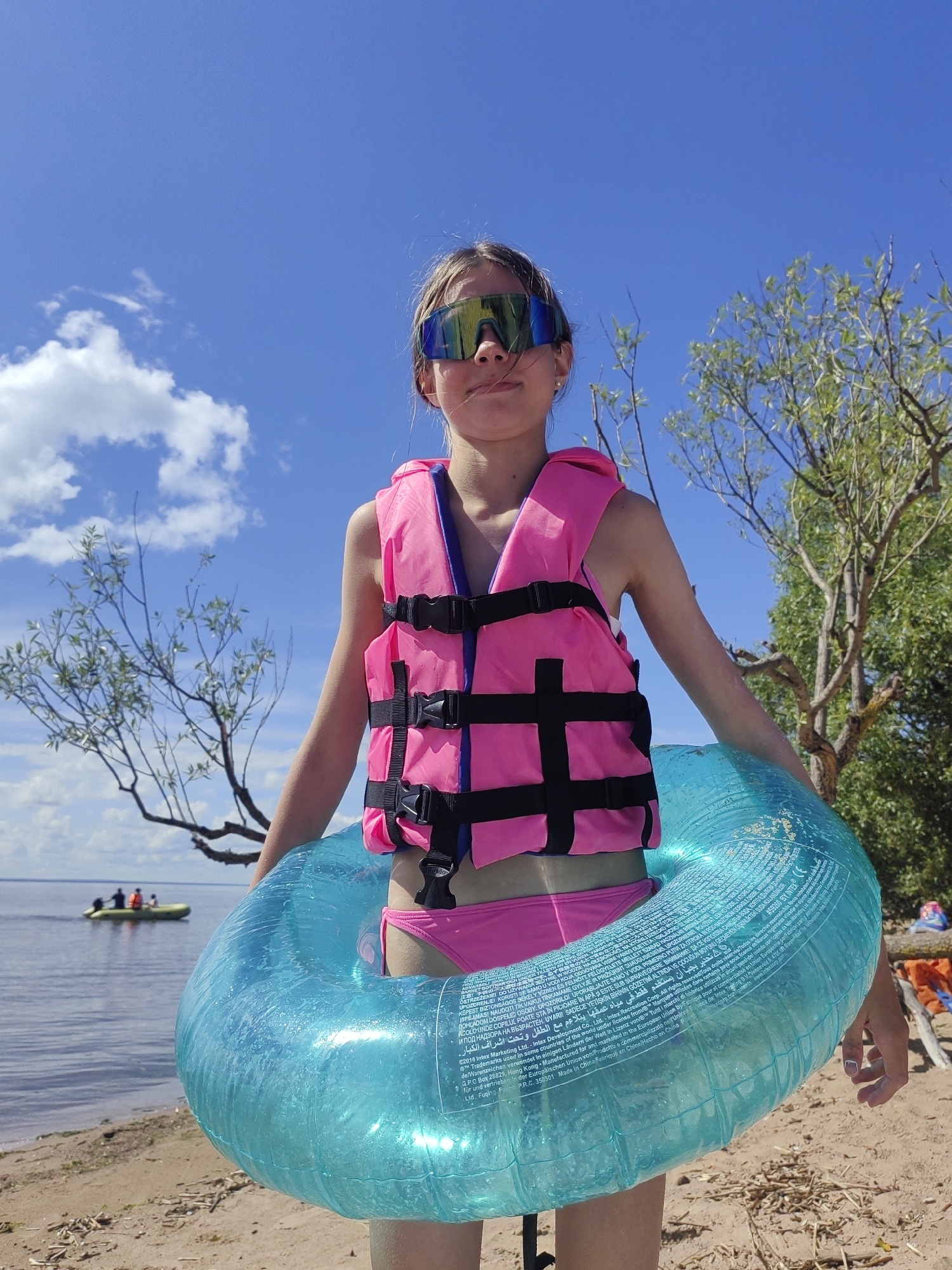 The image shows a young girl standing on a sandy beach, holding a large, transparent blue inflatable tube. She is wearing a bright pink life jacket over a pink swimsuit and oversized mirrored sunglasses. Her hair is light brown and pulled back. The sky above is a clear blue with scattered white clouds. To the left, the calm water of a large lake or bay stretches out, with a small boat carrying several people in the distance. On the right side of the image, there are trees with green foliage and the beach has some scattered driftwood. The scene appears to be set on a sunny day, likely during the summer. The girl is looking directly at the camera with a neutral expression. The inflatable tube has text printed on it in multiple languages, indicating it is a product of Intex. The location context provided is Saint Petersburg, Russia.