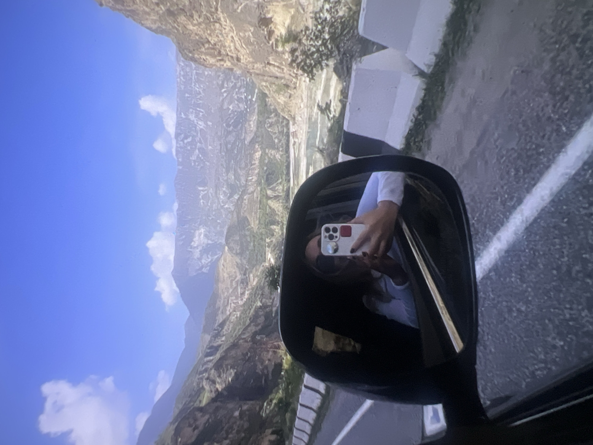The image captures a vibrant mountainous landscape and a self-portrait taken via a car's side mirror. The main subject is a person, likely female, whose reflection is visible in the car's side mirror. They are holding a white smartphone with a prominent camera array (three lenses) in their right hand, angled to take a picture of themselves in the mirror. The person is wearing a light-colored top, and part of their face, dark hair, and an arm are visible.
The background, seen both directly and reflected, is a vast mountain range with steep, rocky slopes. Some parts of the mountains show sparse vegetation, while others appear barren and rugged. The sky above is a clear, bright blue with a few scattered white, fluffy clouds. A paved road runs alongside the mountains, marked by a white concrete barrier or guardrail. The perspective suggests the viewer is inside a vehicle, and the mirror reflects both the person and the passing scenery.
The image was taken during daytime, under clear, sunny weather conditions, which contributes to the sharp visibility of the landscape and the mirror's reflection. There is no discernible text visible in the image. The scene conveys a sense of travel and appreciation for natural scenery.