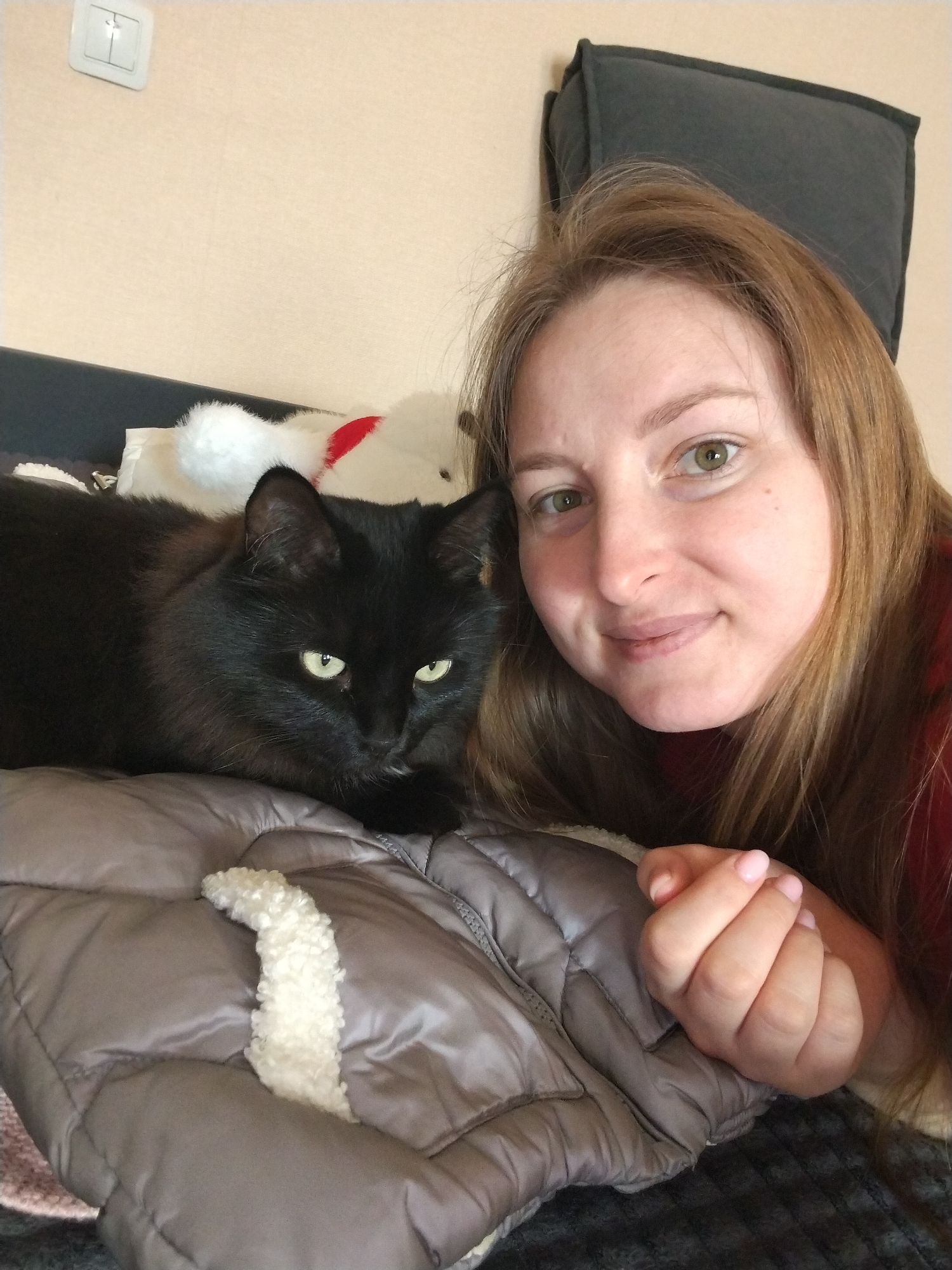 An indoor scene in Chaikovsky, Russia, depicts a Caucasian woman and a black cat lying in close proximity. The woman, with light brown hair and green-hazel eyes, smiles faintly towards the camera. She is partially clothed in a red garment, and her right hand, with light pink nail polish, rests on a taupe-colored padded, quilted jacket or blanket that features a white, curly fleece lining. To her left, a black cat with prominent green-yellow eyes is positioned on its stomach, looking slightly to the left of the camera. Behind the cat, a white plush object with a red accent is partially visible. The background consists of a light beige textured wall, with a white rectangular dual-toggle light switch mounted in the upper left corner. A dark grey cushion is visible behind the woman's head, resting against the wall.