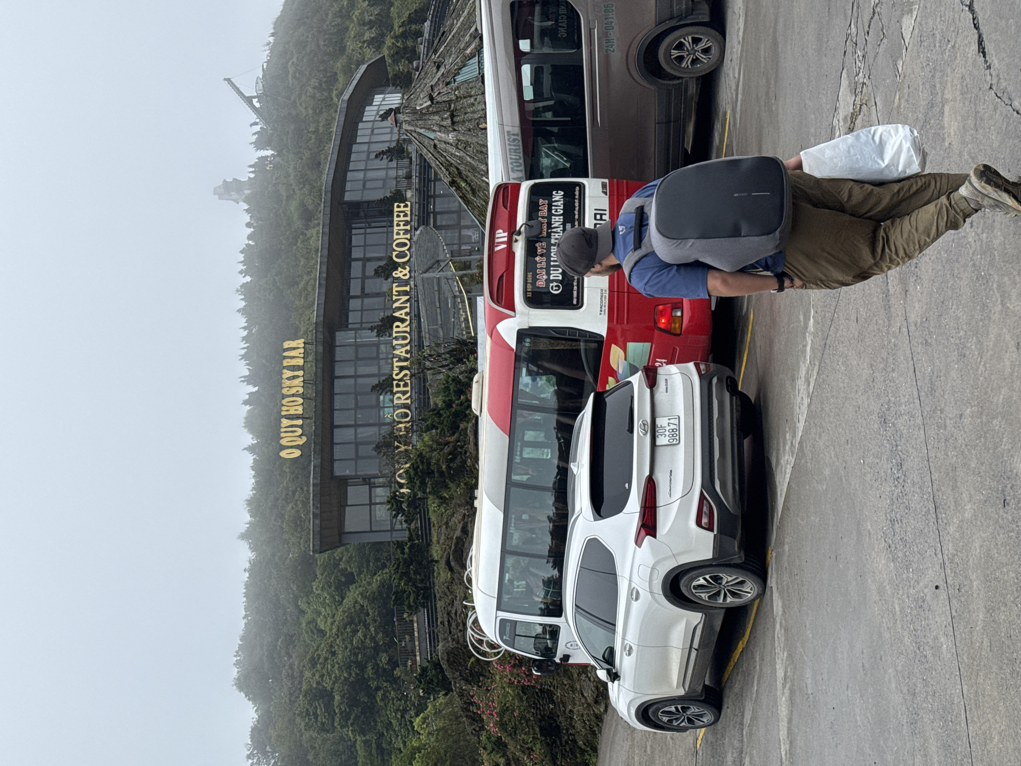 The image shows a street scene in Binh Lu, Vietnam, with a white Hyundai SUV displaying the license plate "30F 988.71" parked along the curb. A person wearing a backpack and holding a white bag walks along the sidewalk. A red and white bus with the words "A TOURIST" and "XE HOP DONG" is parked next to the car. Behind the bus is a brown van. Visible in the background is a building with the words "O QUY HO SKY BAR" and "RESTAURANT & COFFEE" on its facade. The landscape is mountainous and covered with trees, and the sky is overcast.